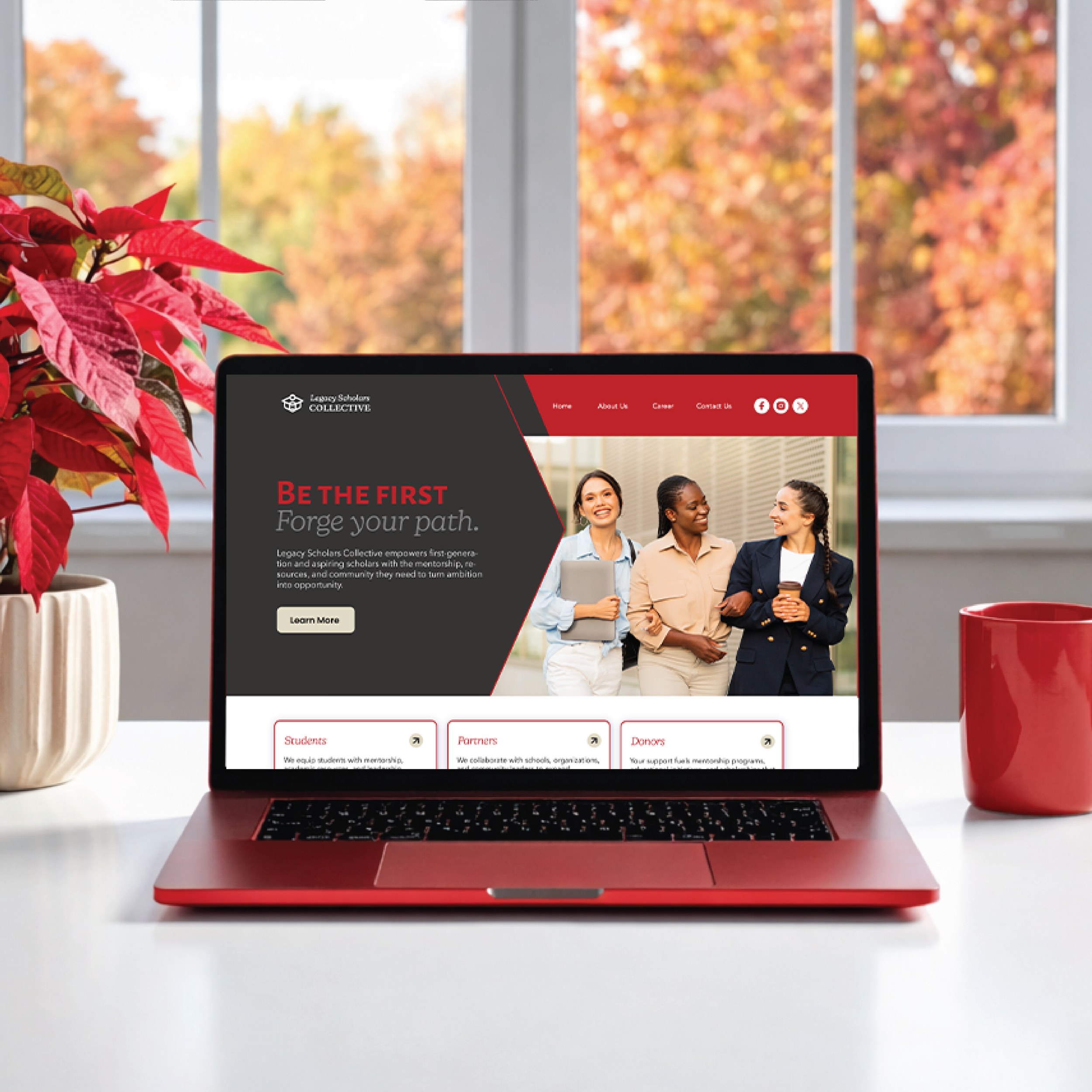Close-up of a red laptop on a white surface with a website page displaying a group of diverse women talking, a poinsettia plant on the left, and a window with fall foliage visible outside.