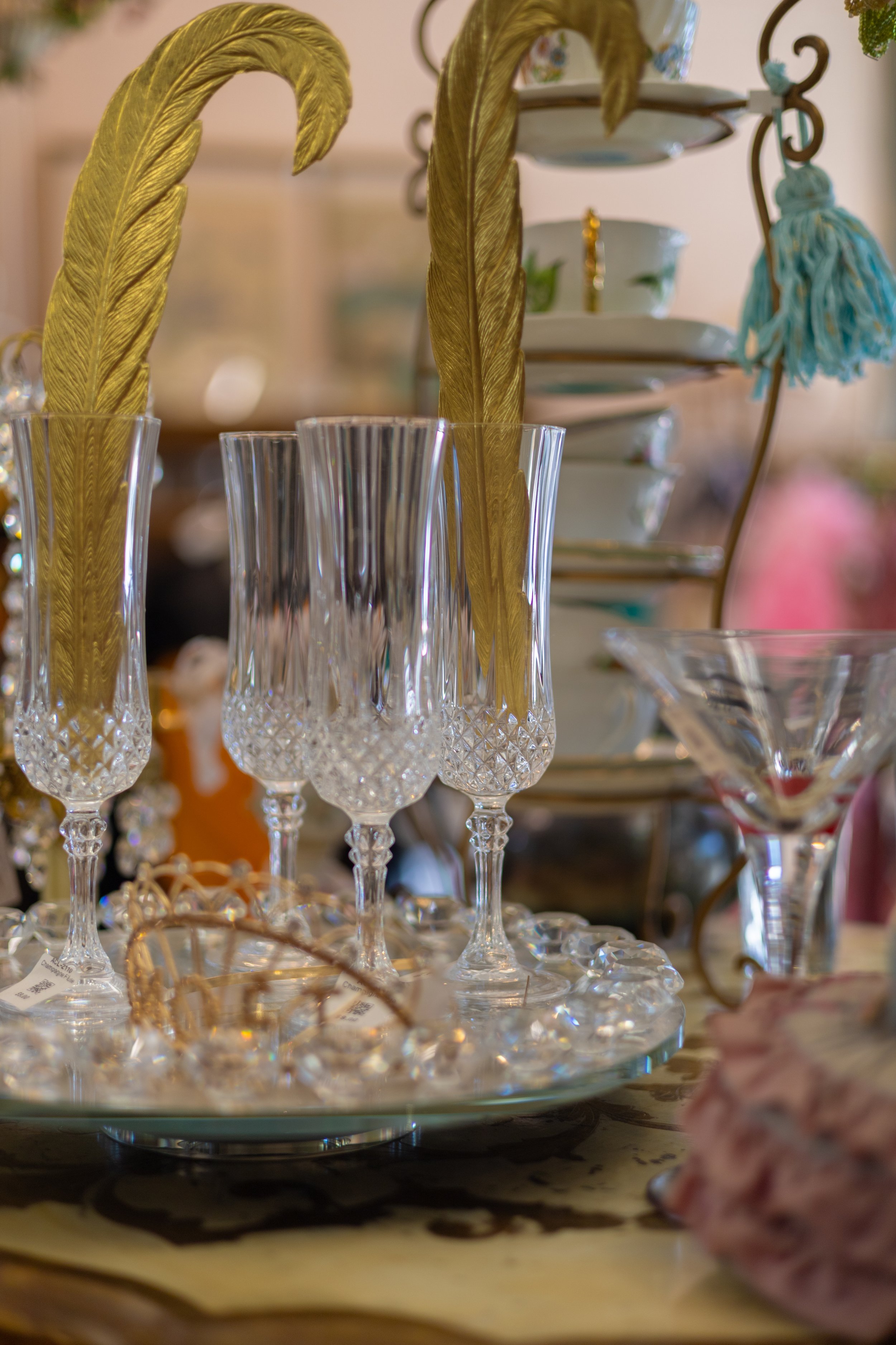 A display of clear and gold decorative glassware, including tall champagne flutes with intricate patterns, along with tiered serving plates in the background, set on a table with a blurred backdrop.