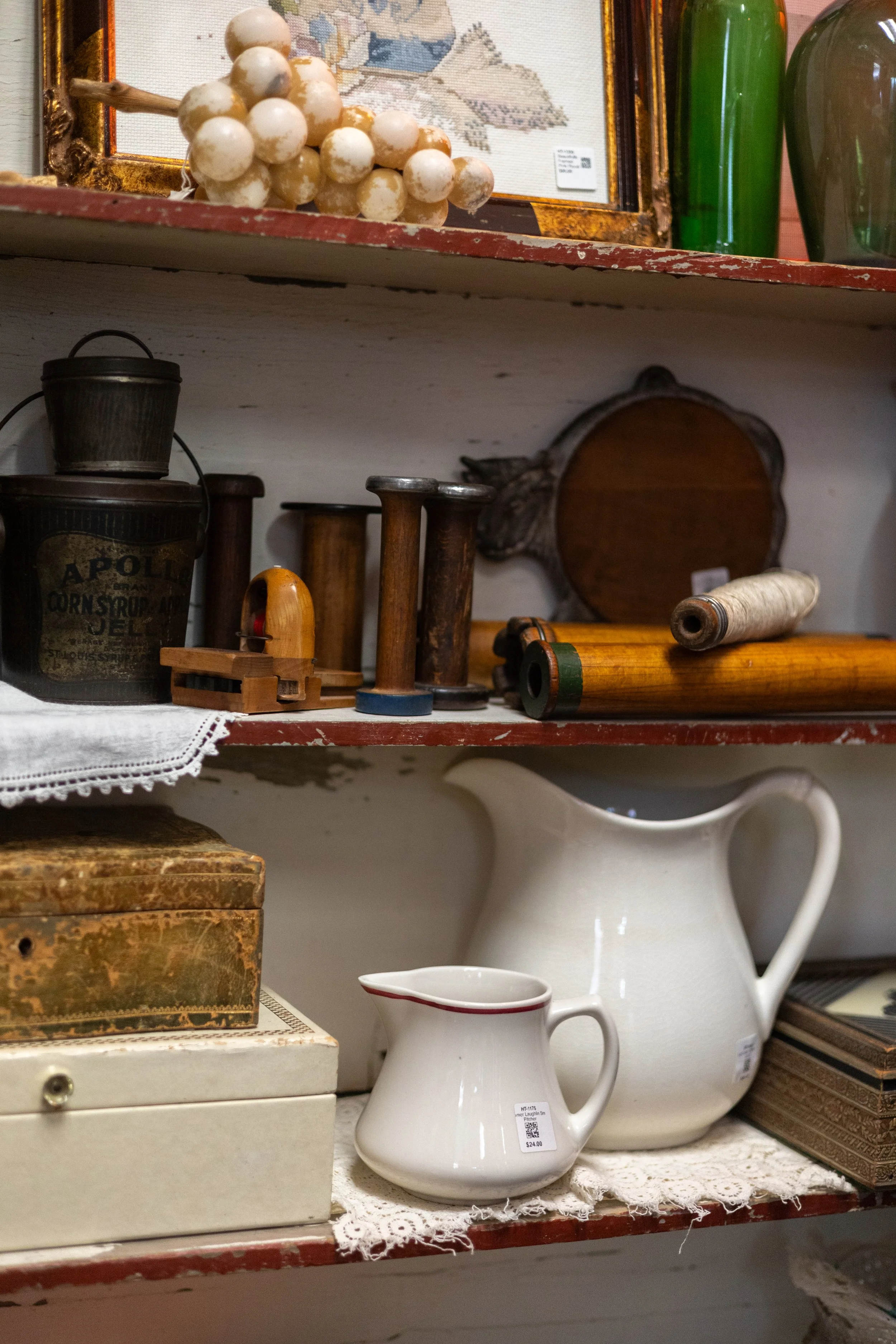 Various vintage and antique household items on a shelf, including a bunch of dusty white grapes, old spools of thread, a small box, a white ceramic pitcher, and a framed cross-stitch artwork.
