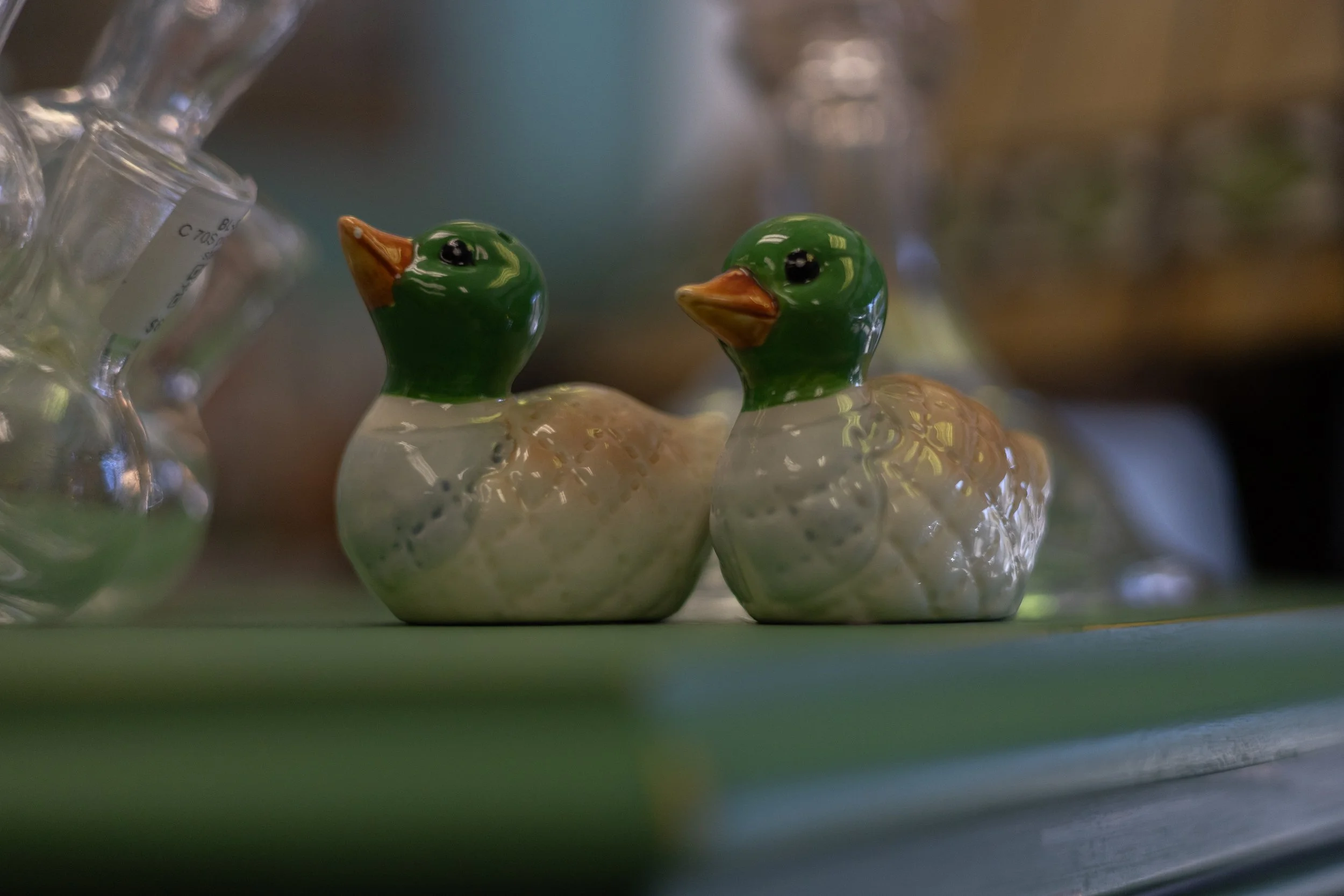 Two ceramic duck figurines, one with a green head and one with a beige head, are displayed on a shelf with blurred glassware in the background.