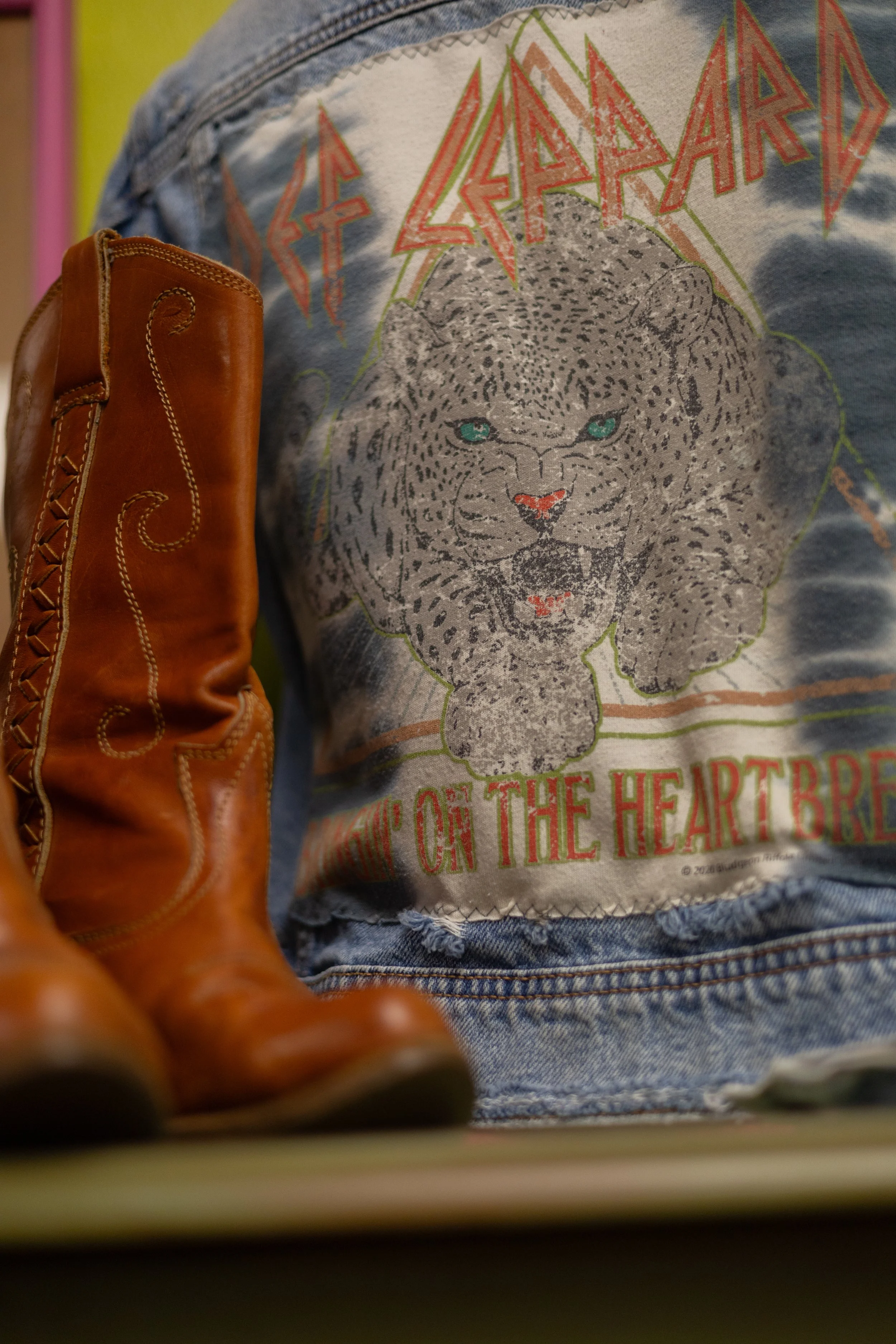 Close-up of a person wearing a distressed denim jacket with a graphic of a leopard's face and colorful text, with a brown leather boot partially visible in the foreground.