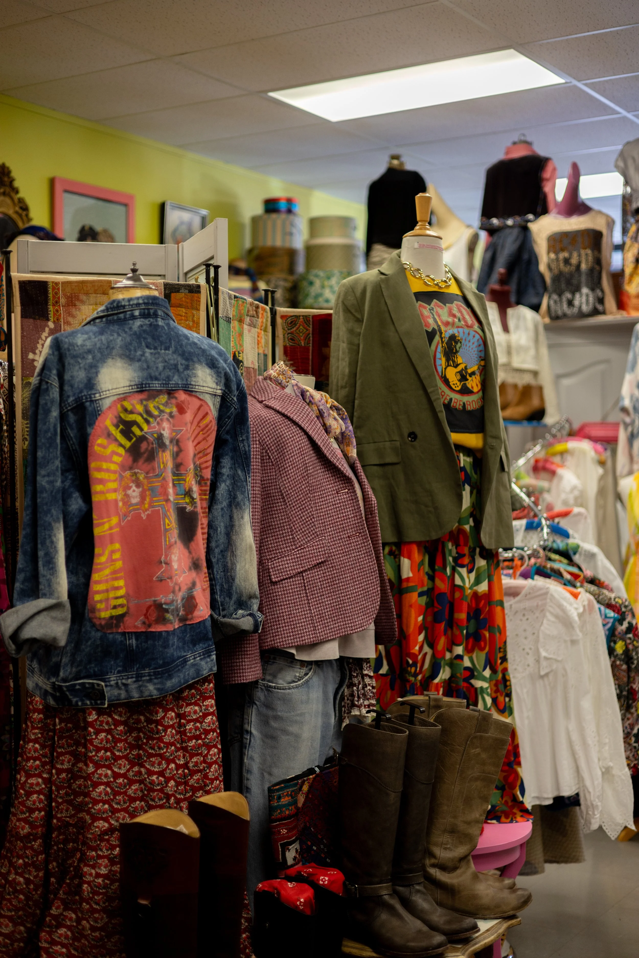 Clothing display in a thrift store, including denim jackets with colorful patches, a pink blazer, patterned skirts, vintage boots, and mannequins dressed in casual and vintage-style outfits in a boutique setting.