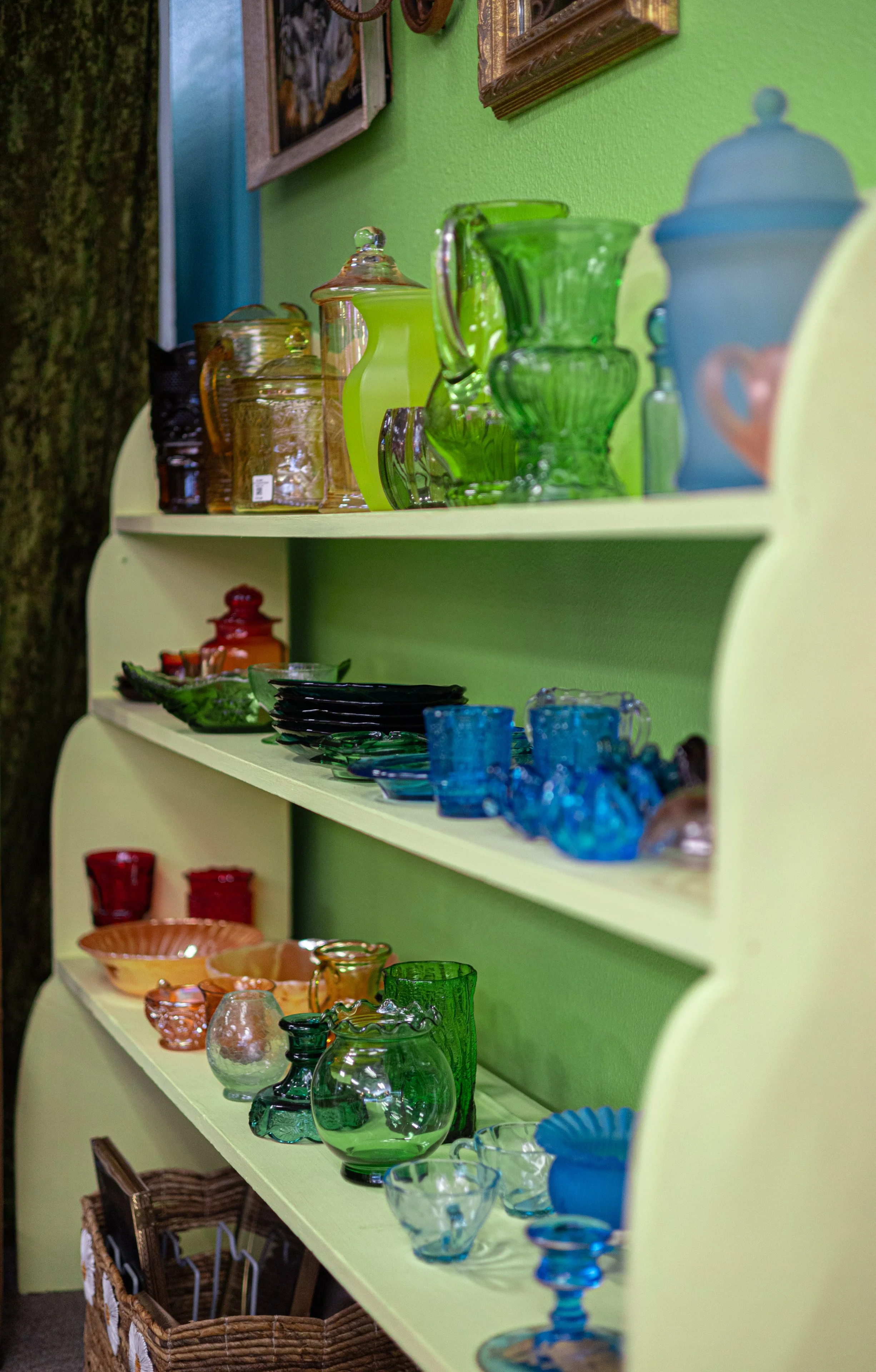 Colorful glassware including jars, pitchers, bowls, and cups arranged on a pastel green shelf with framed artwork and a green wall in the background.