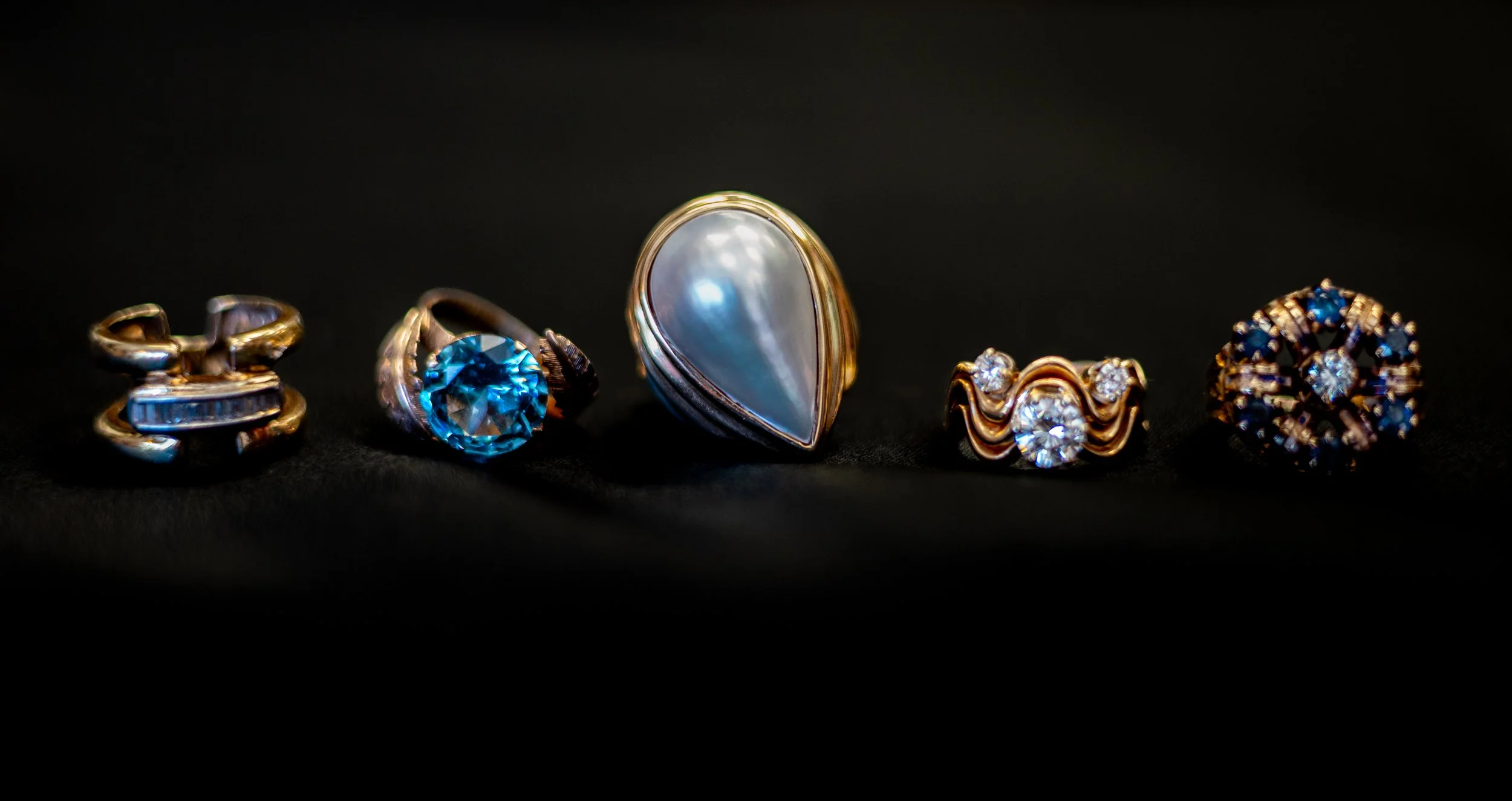 Five rings with various gemstones and designs, including a large pearl ring, displayed on a black background.