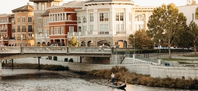 A gondola on a river with two people, with a cityscape featuring colorful buildings and trees in the background.
