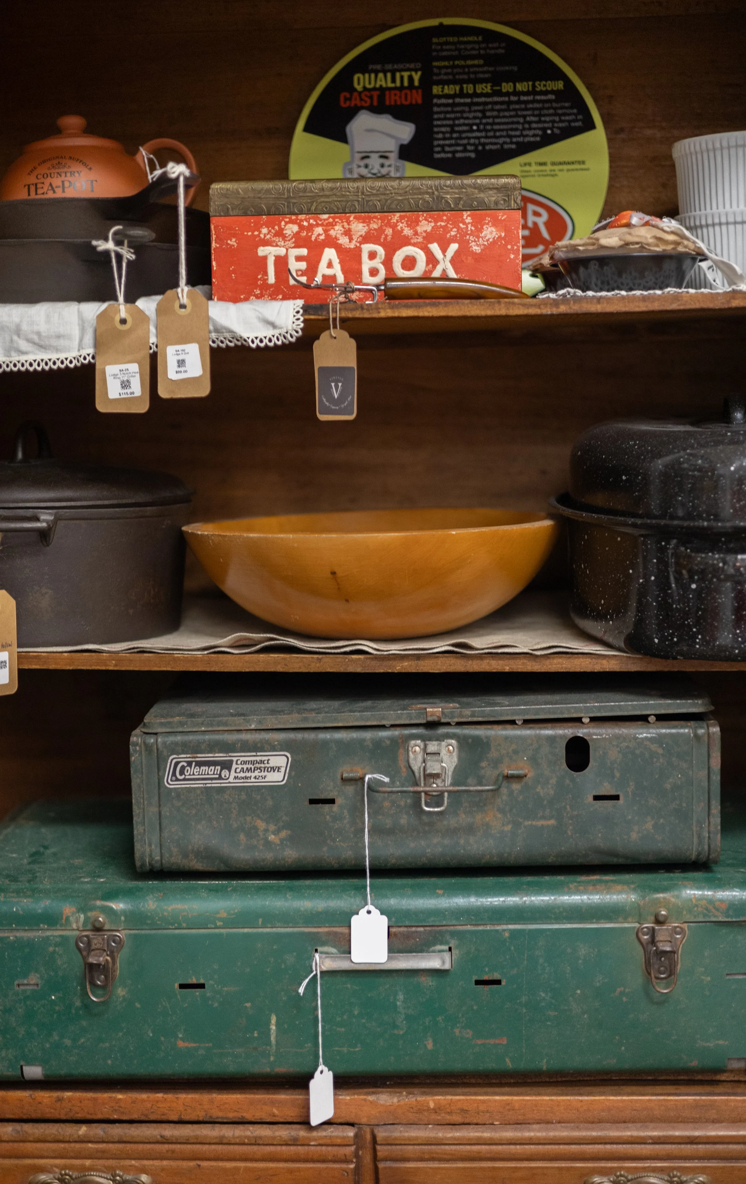 Various vintage kitchen items and containers on shelves, including a red tin labeled 'TEA BOX', a black cast iron pot, a yellow bowl, an orange teapot labeled 'COUNTRY TEA-POT', and a green Coleman stovetop beneath, with price tags hanging from some 