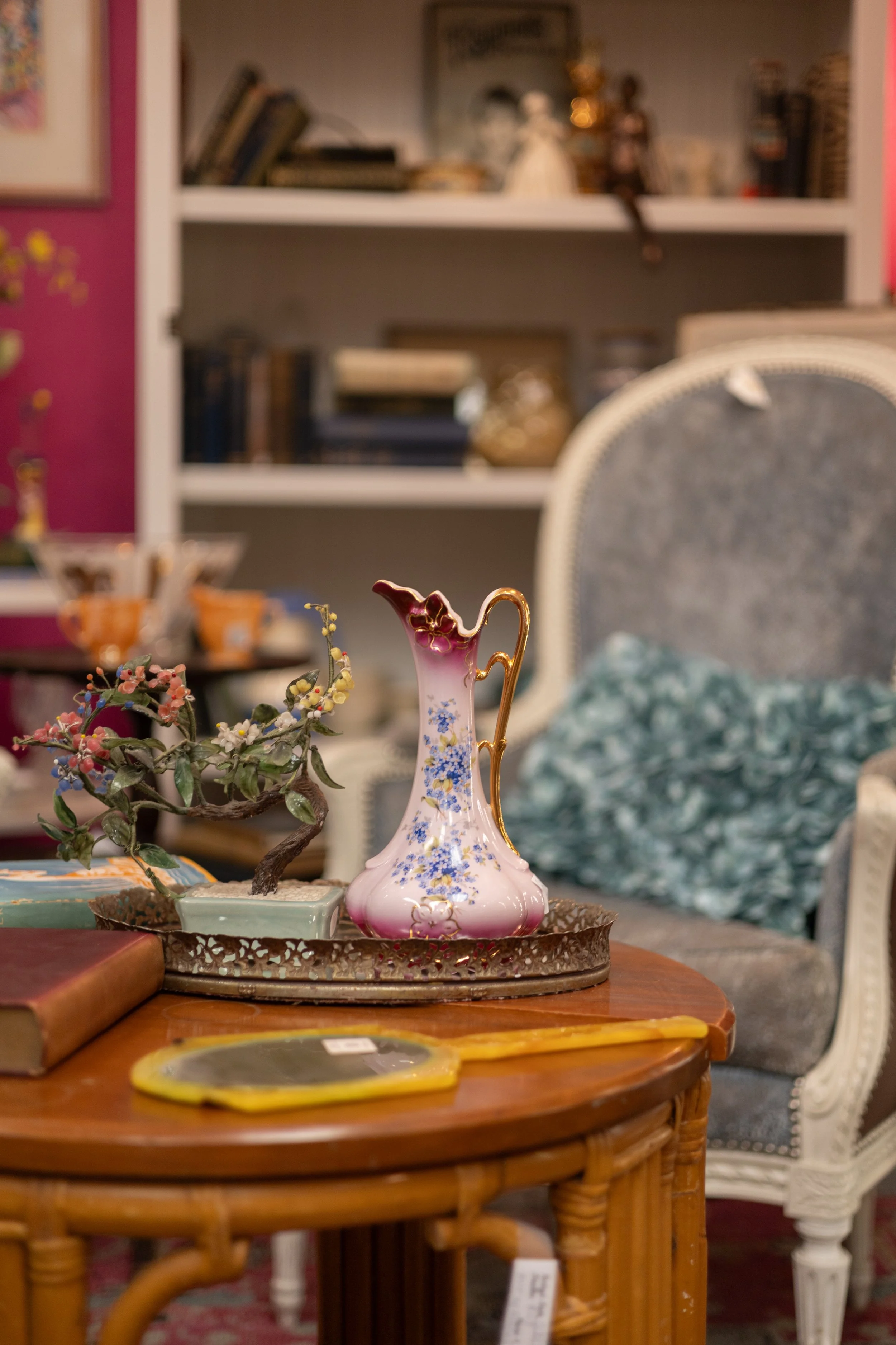 A pink and white decorative porcelain vase with blue floral patterns and gold accents on a metal tray atop a round wicker table, surrounded by other antique furniture and decorative items in a room with bookshelves and upholstered chairs.