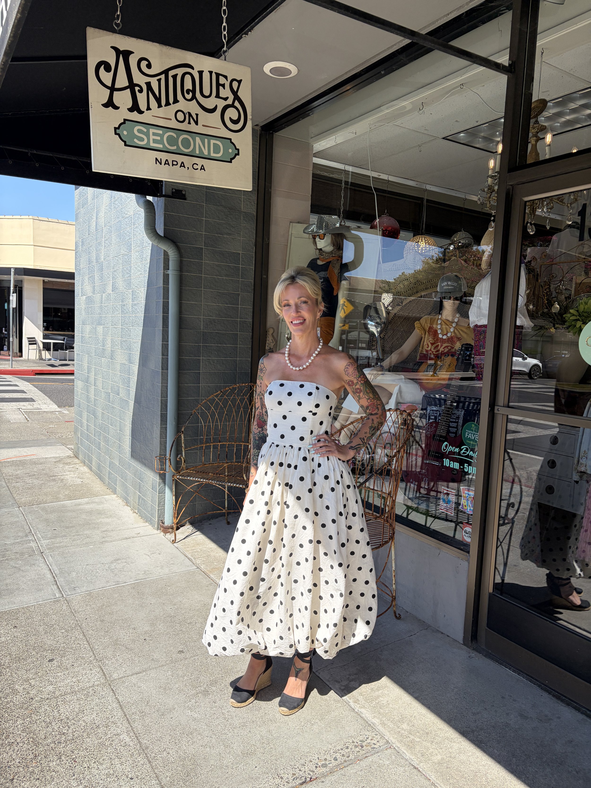A woman standing outside an antique shop called 'Antiques on Second' in Napa, California. She is wearing a vintage-style white dress with black polka dots, black wedge heels, and a pearl necklace. She has floral tattoos on her arms and is smiling at the camera.