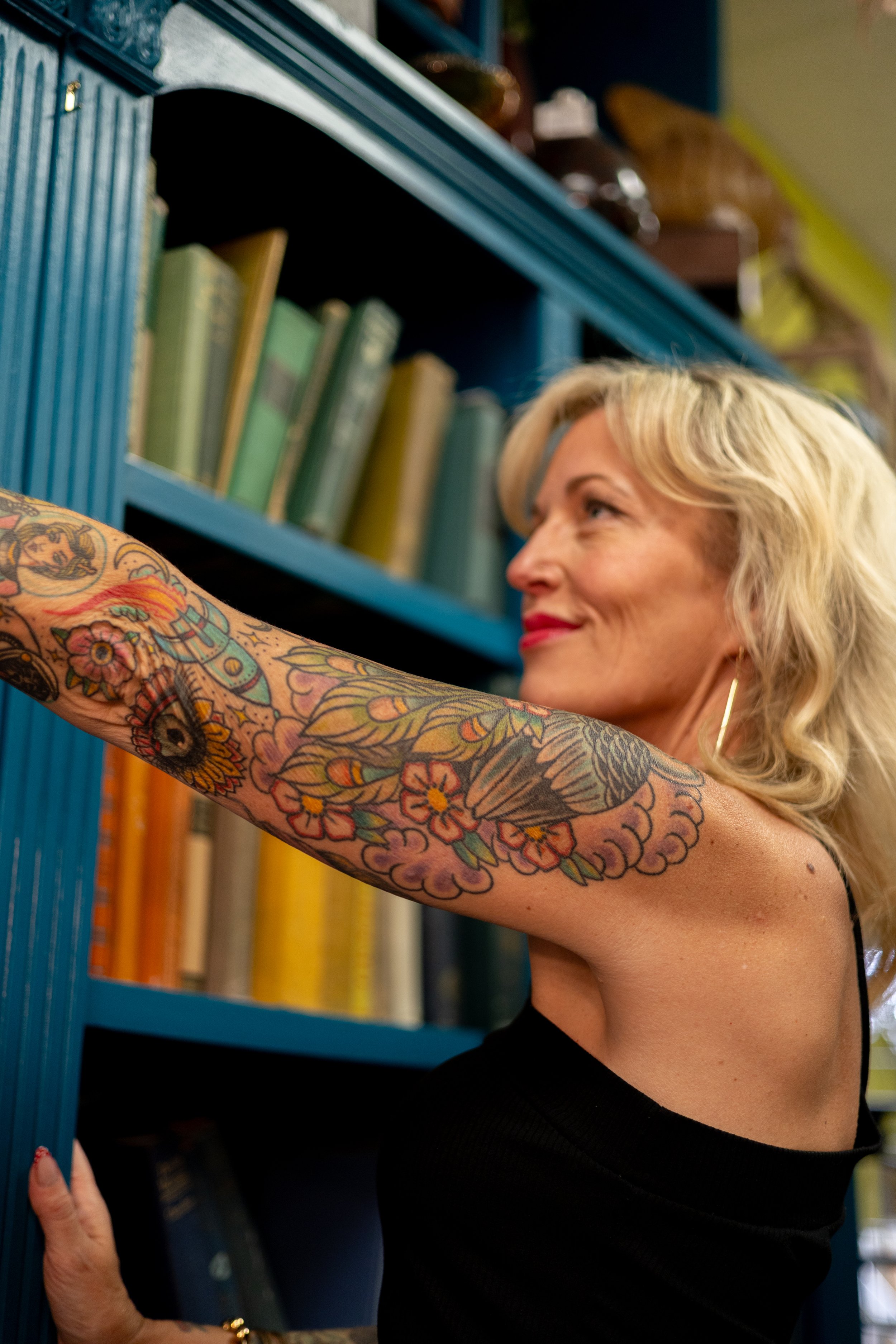 A woman with blonde hair and colorful tattoos on her arm reaching to the top shelf of a blue bookshelf filled with books.