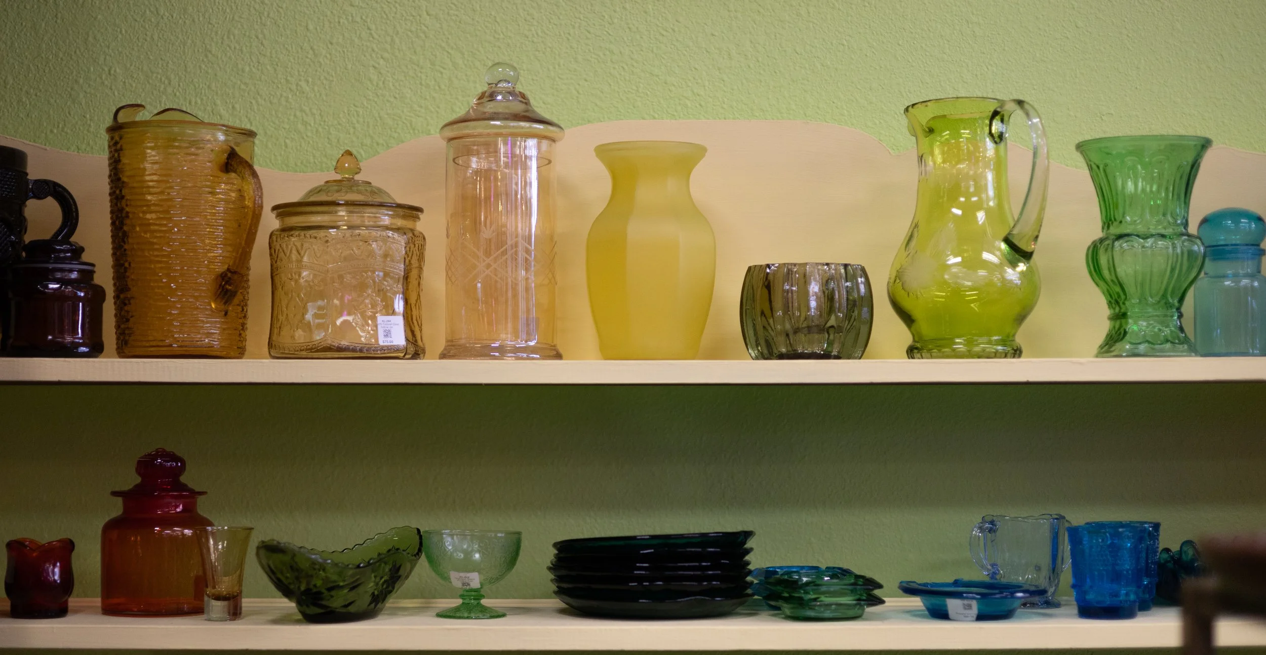 A collection of colorful glassware including vases, bowls, and pitchers arranged on shelves against a green wall.