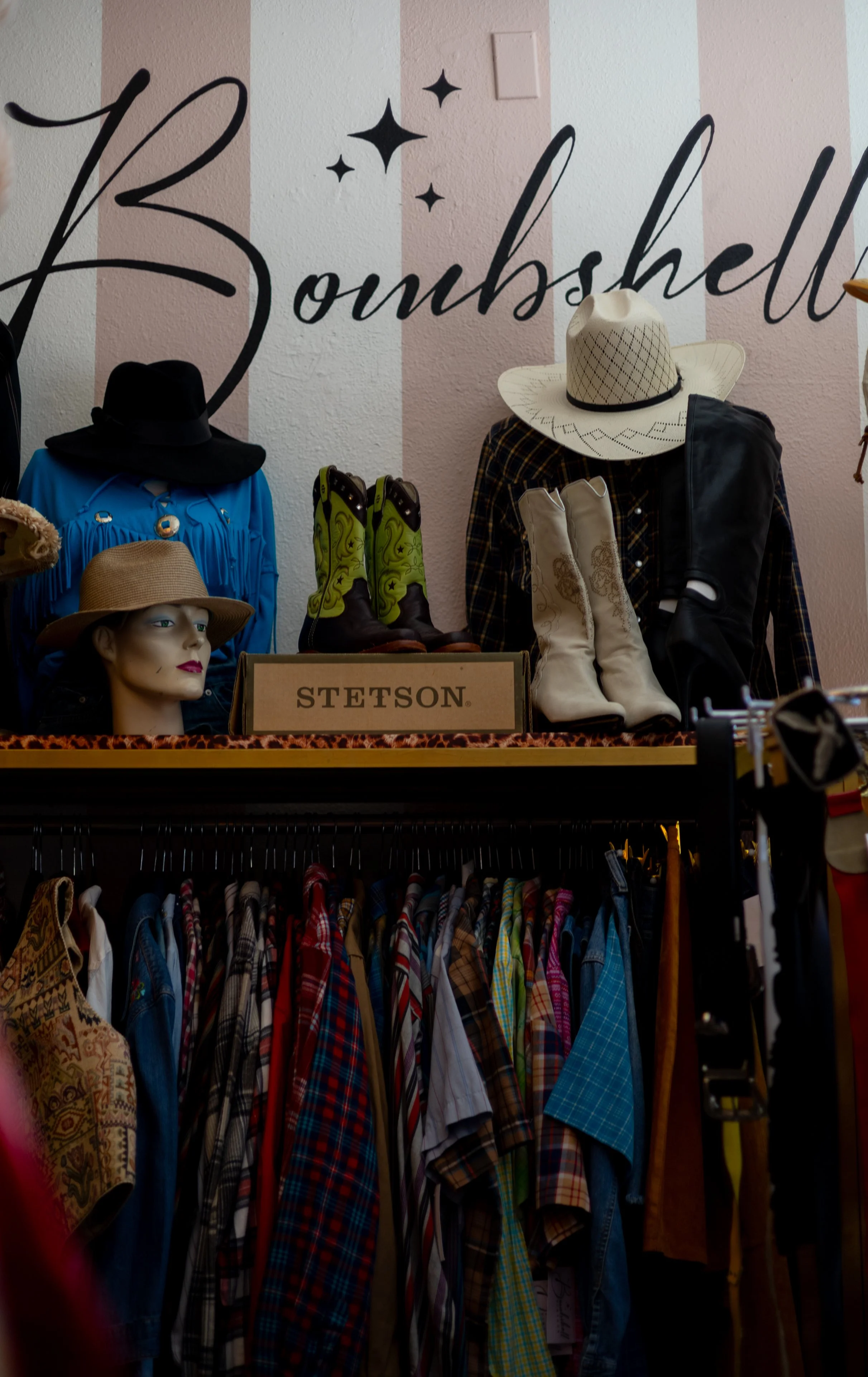 Display of vintage and Western clothing and boots, including a mannequin head with a hat, plaid shirts, and colorful cowboy boots, in a thrift shop aisle.