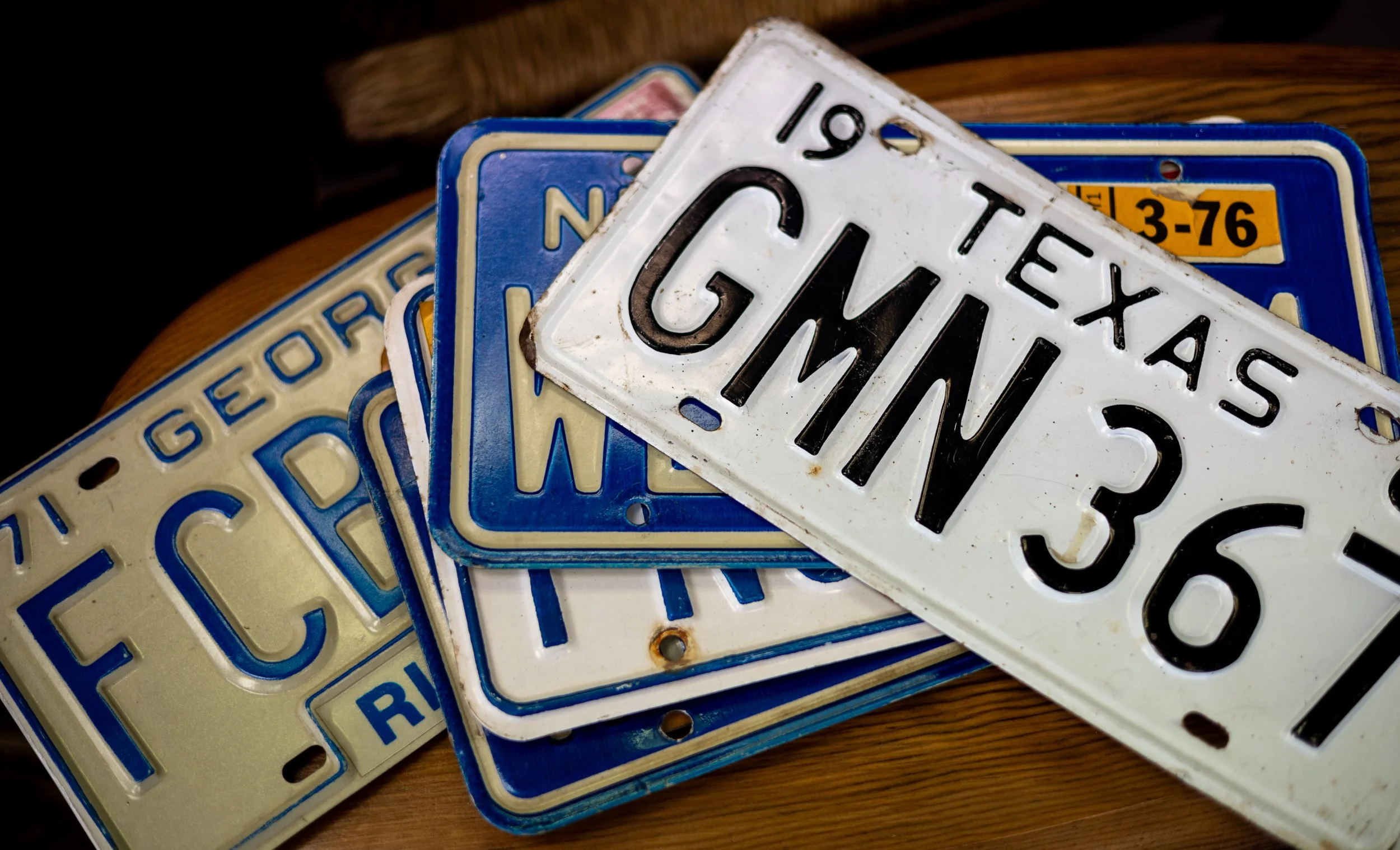 A pile of vintage license plates, including a white one with the word 'GWAIT' and other plates with blue and cream colors, all stacked on a wooden surface.