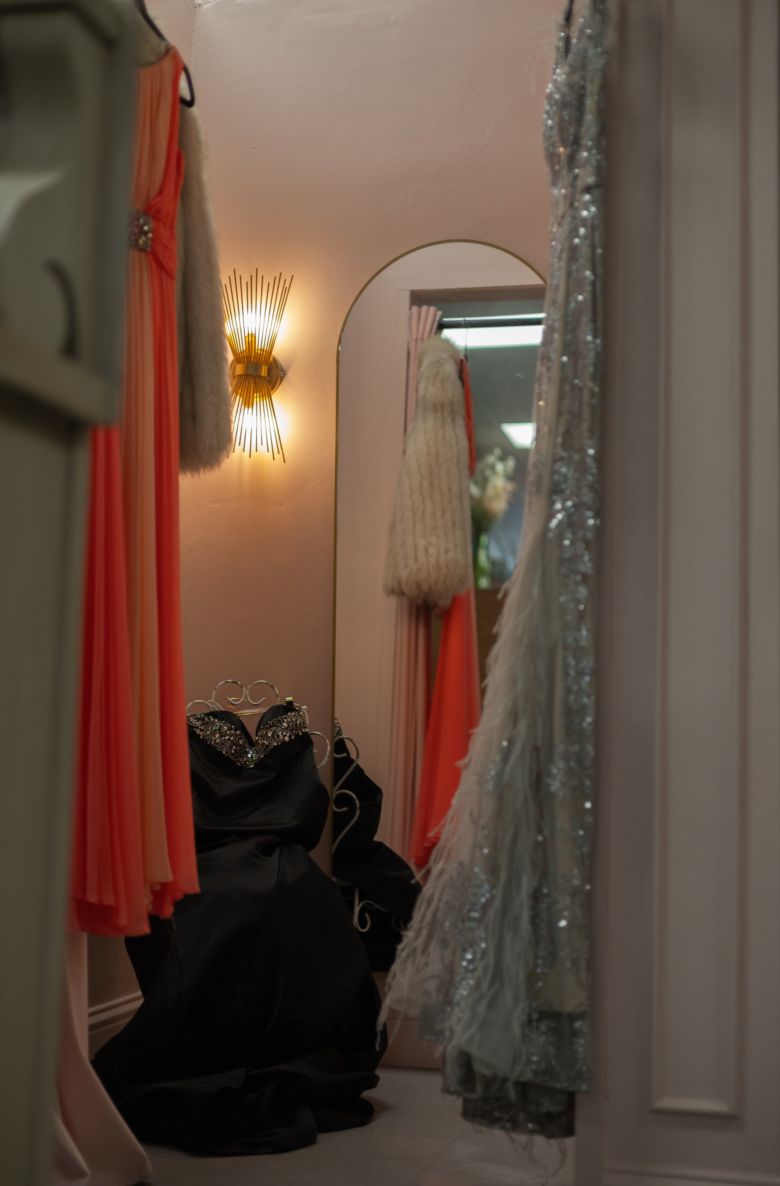 A fitting room with various dresses hanging, a mirror reflecting a clothing rack, and a person crouching on the floor surrounded by clothing and shoes.