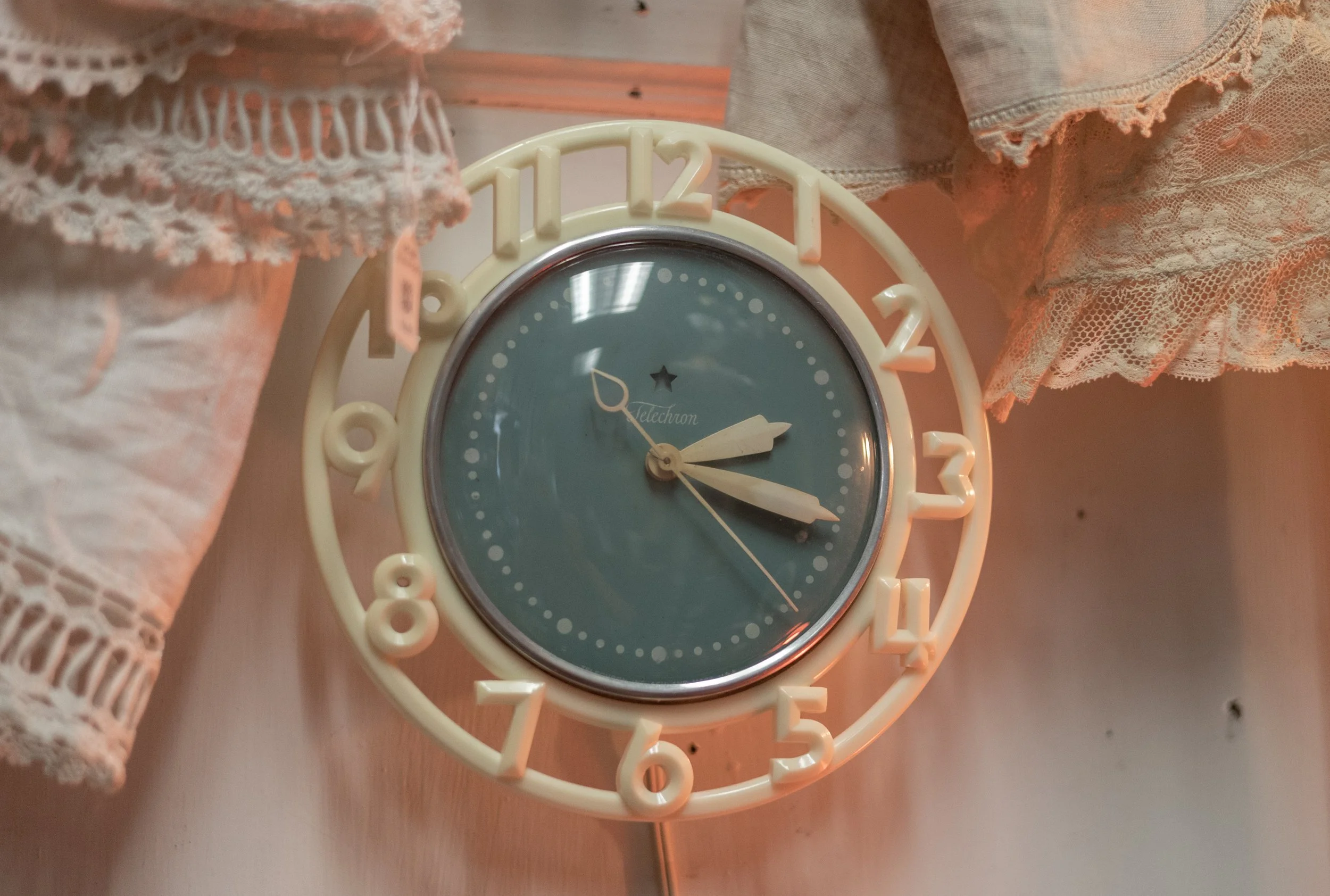 A vintage tabletop clock with a blue face and white numbers, showing the time as 3:20, positioned among pink and beige lace fabric decorations.