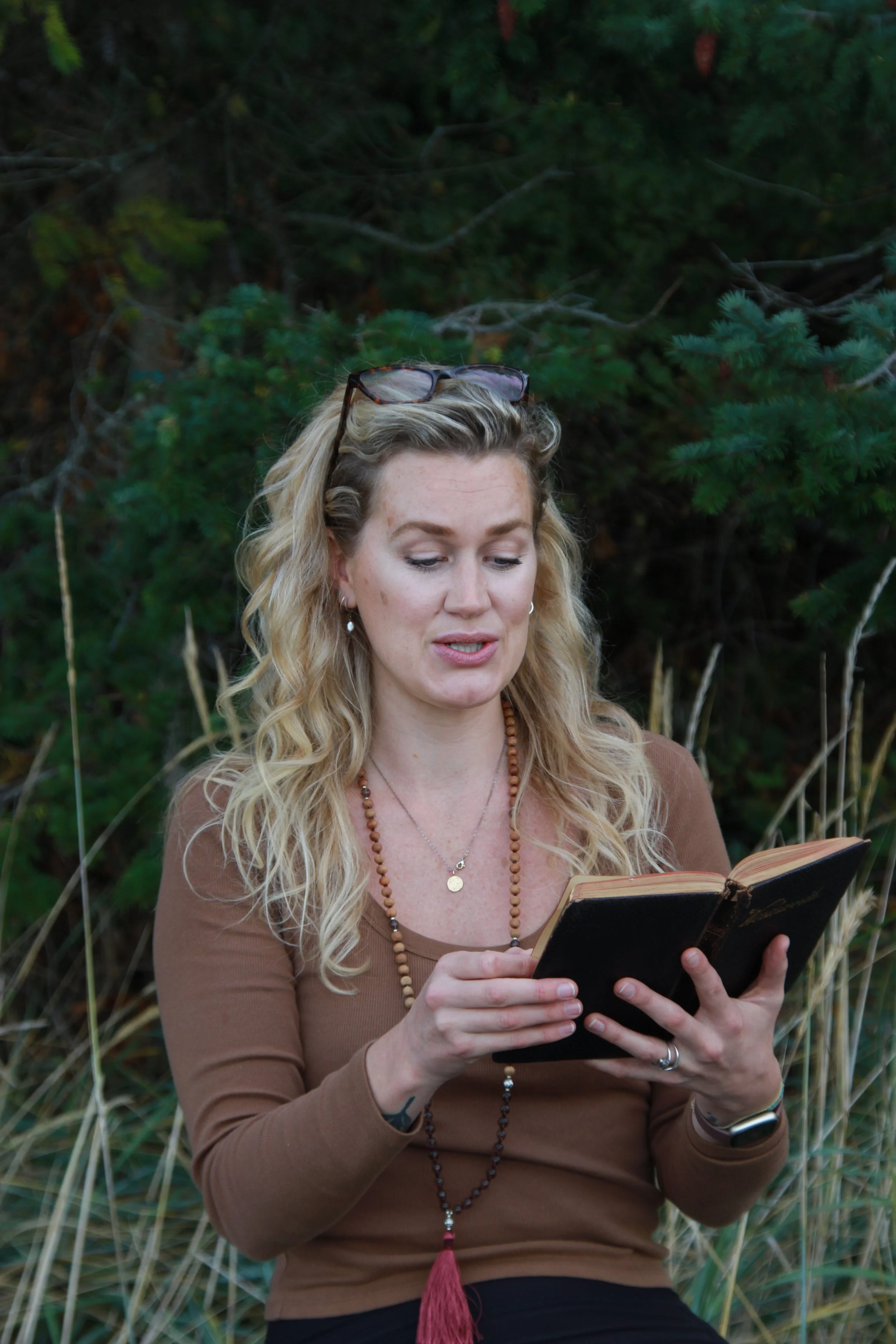 A woman with long curly blonde hair, wearing glasses on her head, a brown long-sleeved top, and jewelry, is reading a book outdoors surrounded by greenery.
