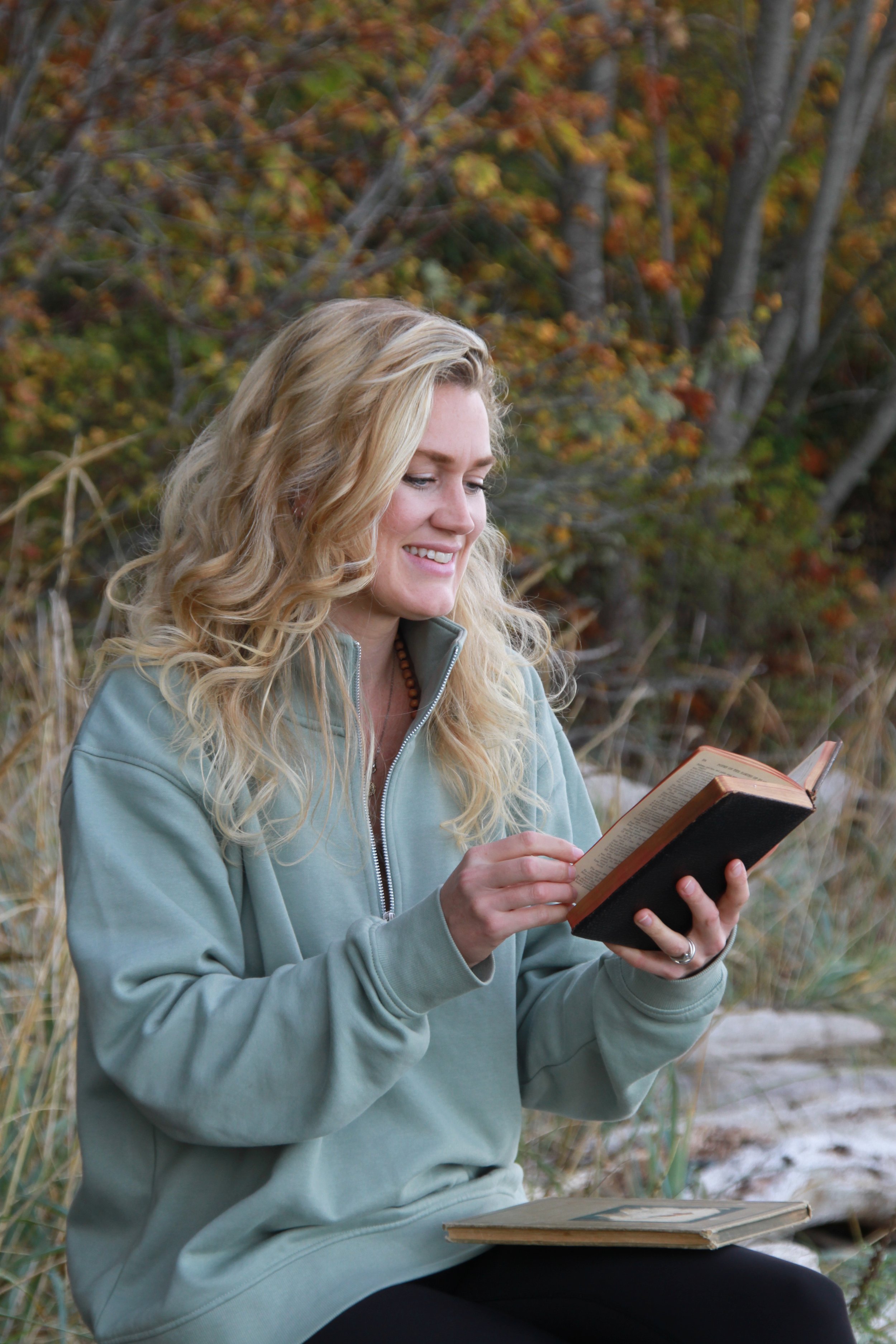 A woman with long blonde wavy hair, wearing a light teal hoodie, sitting outdoors in a natural setting with trees and fall foliage, reading a book and smiling.