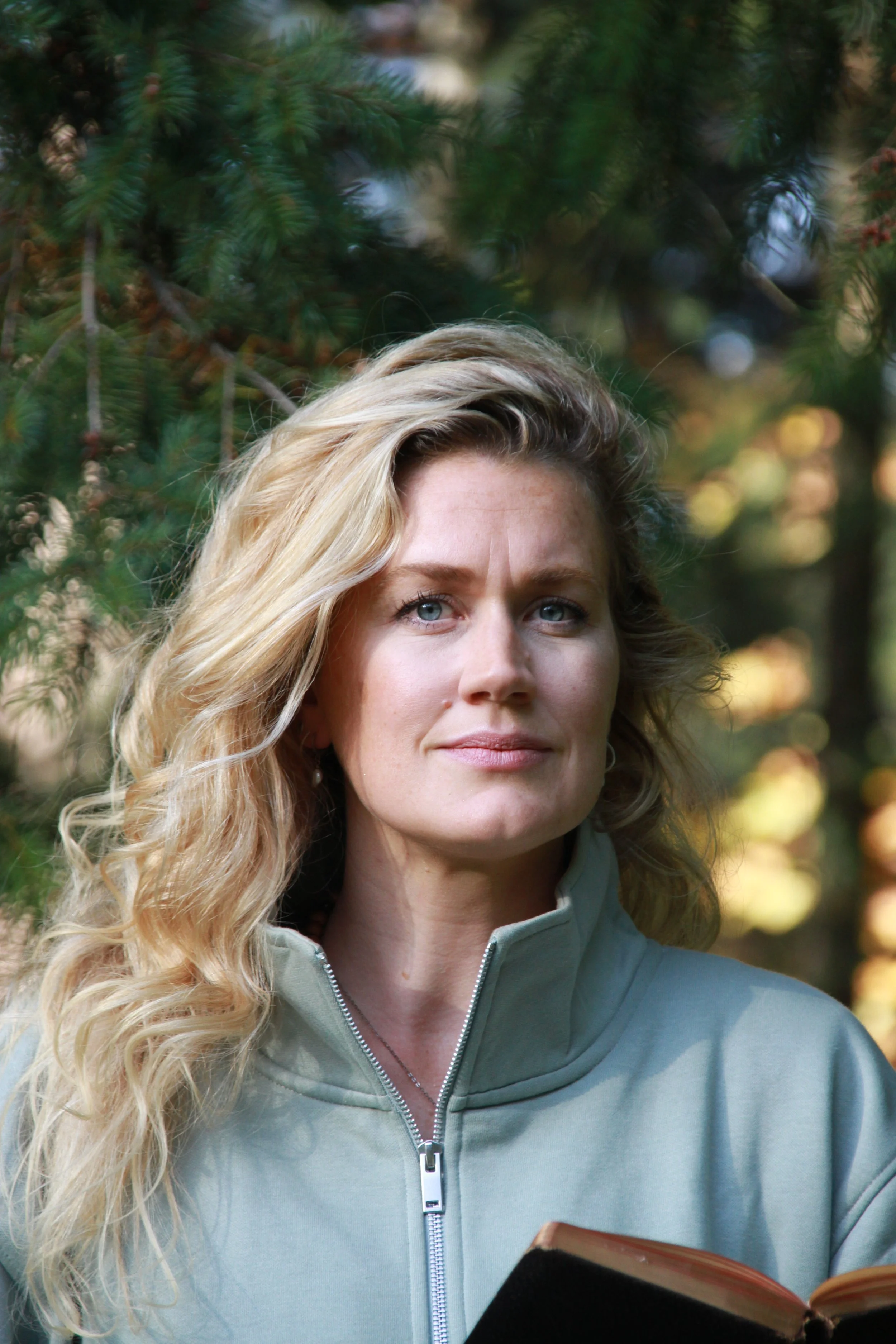 A woman with long, wavy blonde hair is outdoors, surrounded by green pine trees. She is wearing a light gray zip-up jacket and holding an open book.