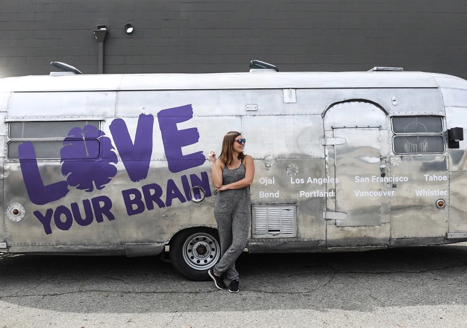 A woman in gray patterned jumpsuit and sunglasses stands and leans against a silver trailer with a large purple 'LOVE YOUR BRAIN' graphic and a list of city names