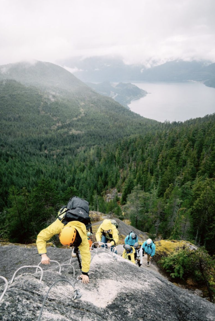 Group of climbers ascending a steep rocky mountain with a forested landscape and a body of water in the background.