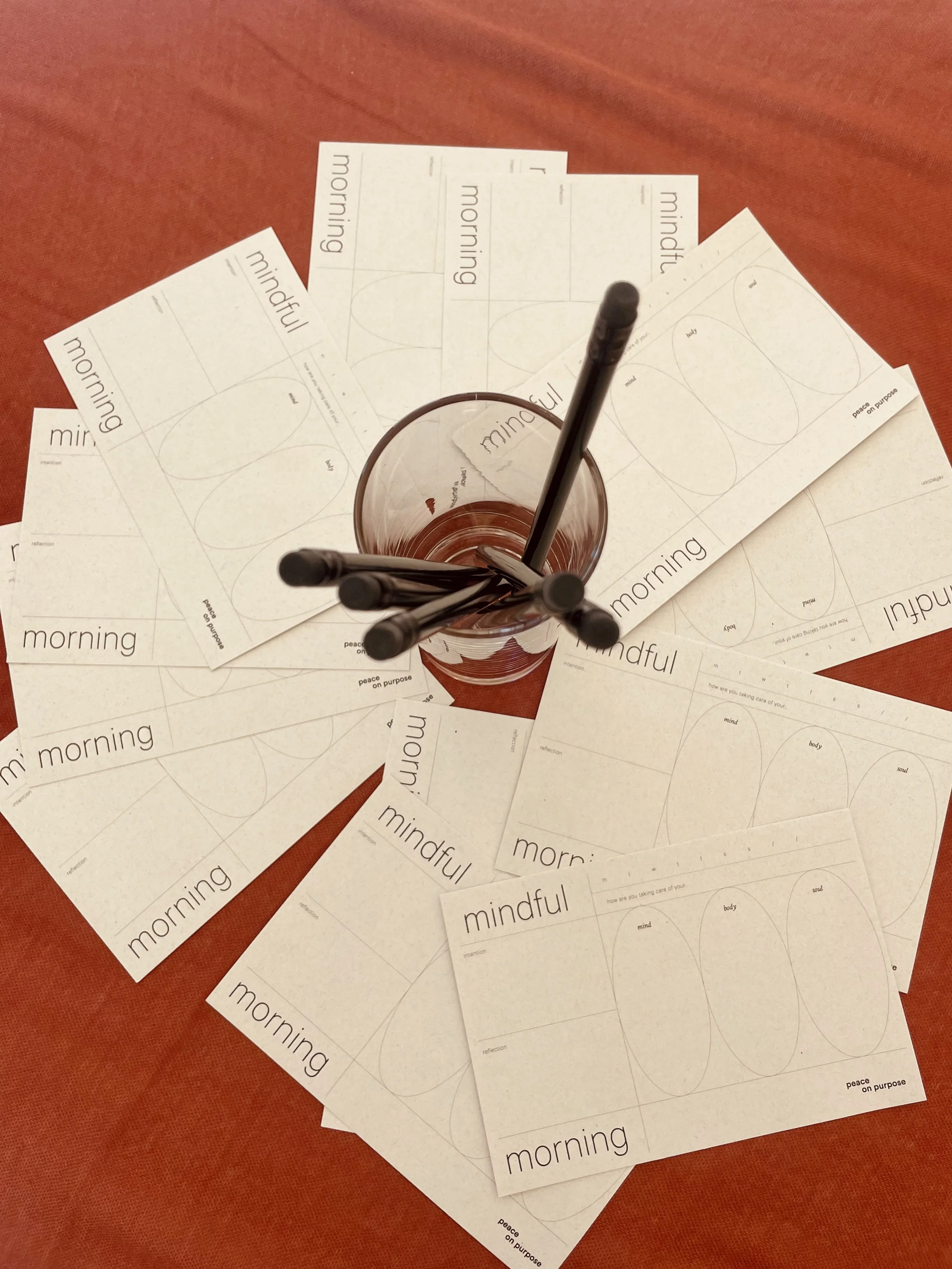 Flat lay of multiple sheets of free printout worksheets with the word 'morning' printed on top, arranged on a reddish-orange surface with a glass in the center holding several black markers.