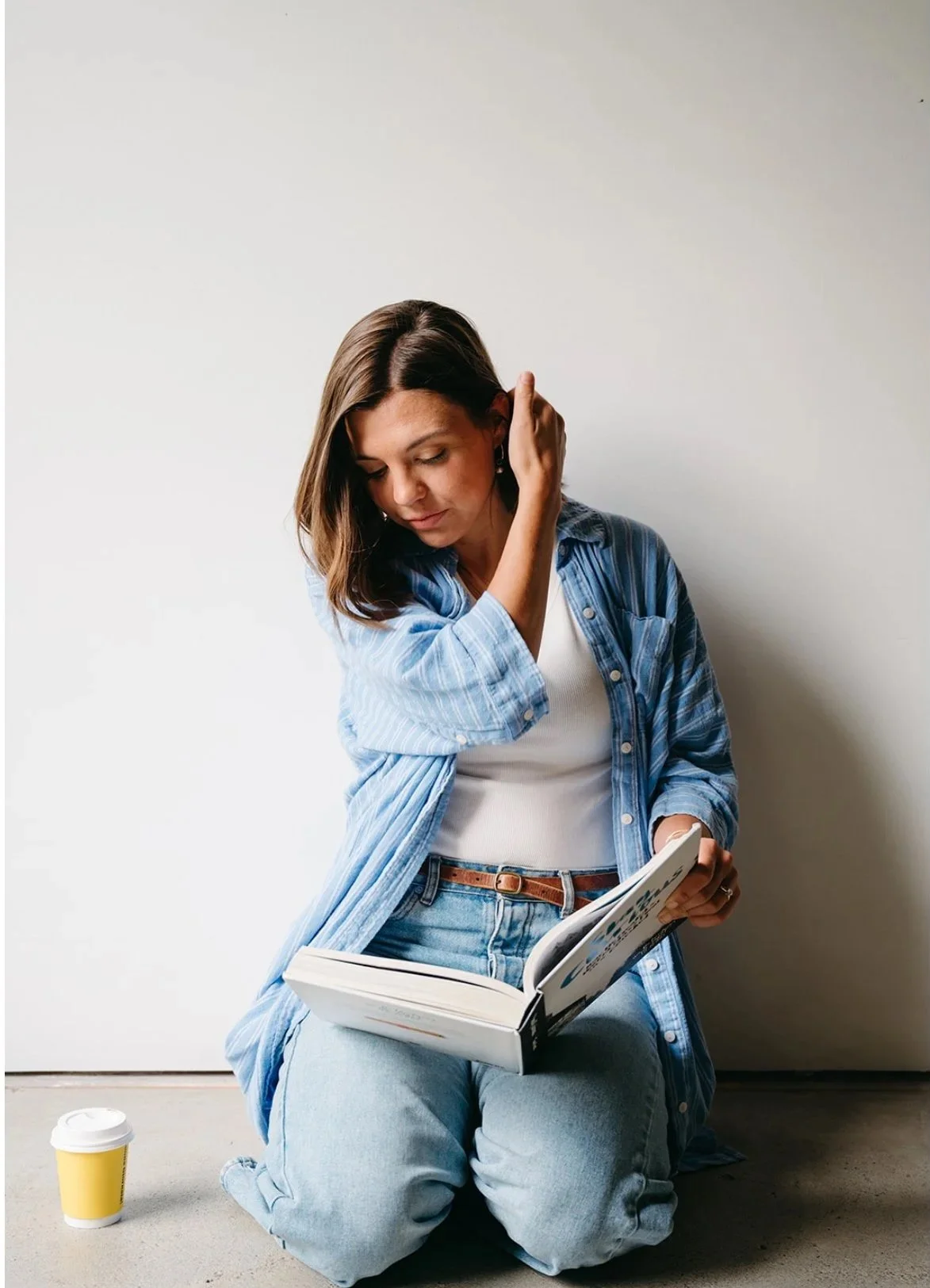 A woman sitting on the floor with a coffee cup nearby, reading a book and touching her ear.