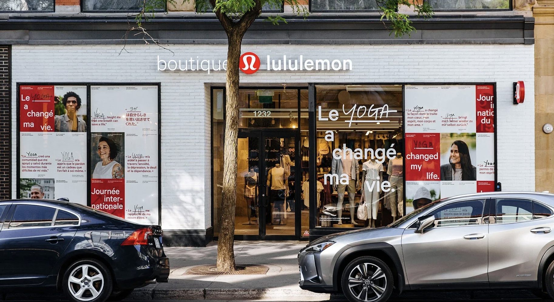 The storefront of a boutique Lululemon store with large windows displaying mannequins dressed in yoga apparel. The store's sign and logo are visible above the entrance, with the words 'boutique Lululemon'. The windows have posters with quotes about yoga and life, featuring images of diverse smiling individuals. Two cars are parked in front of the store, and a small tree partially obscures the view.