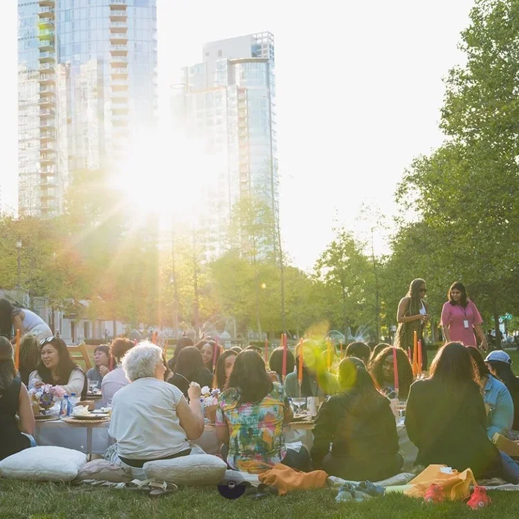 Group of people having a picnic outdoors in a park with tall modern buildings in the background during sunset, some standing and others sitting on cushions.