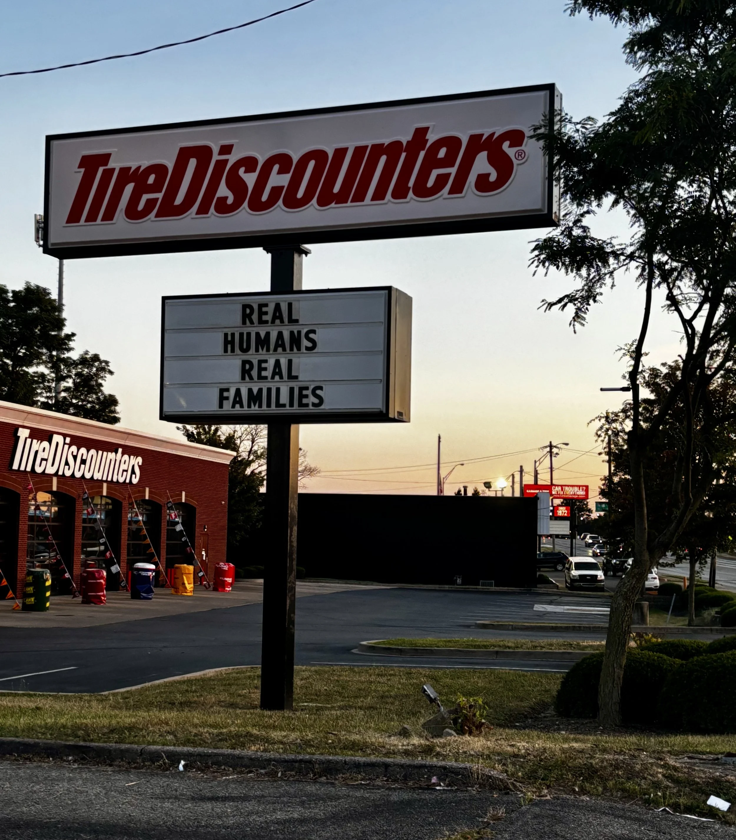 Tire Discount store sign with a message board below it that reads 'Real Humans Deal Real Families' during dusk, with trees, parked cars, and a red brick building in the background.