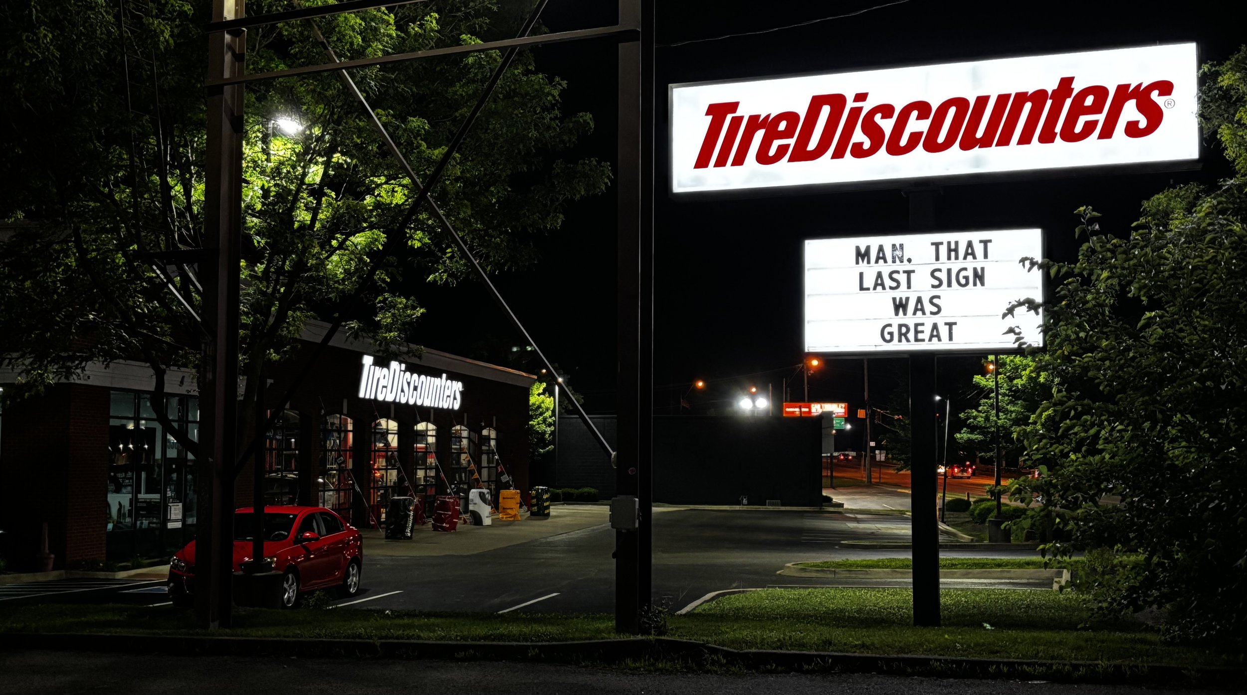 Nighttime view of a TireDiscounters store with illuminated signs, one reading 'TireDiscounters' and another with a message board saying 'MAN. THAT LAST SIGN WAS GREAT'. There are trees, parked cars, and streetlights in the surrounding area.