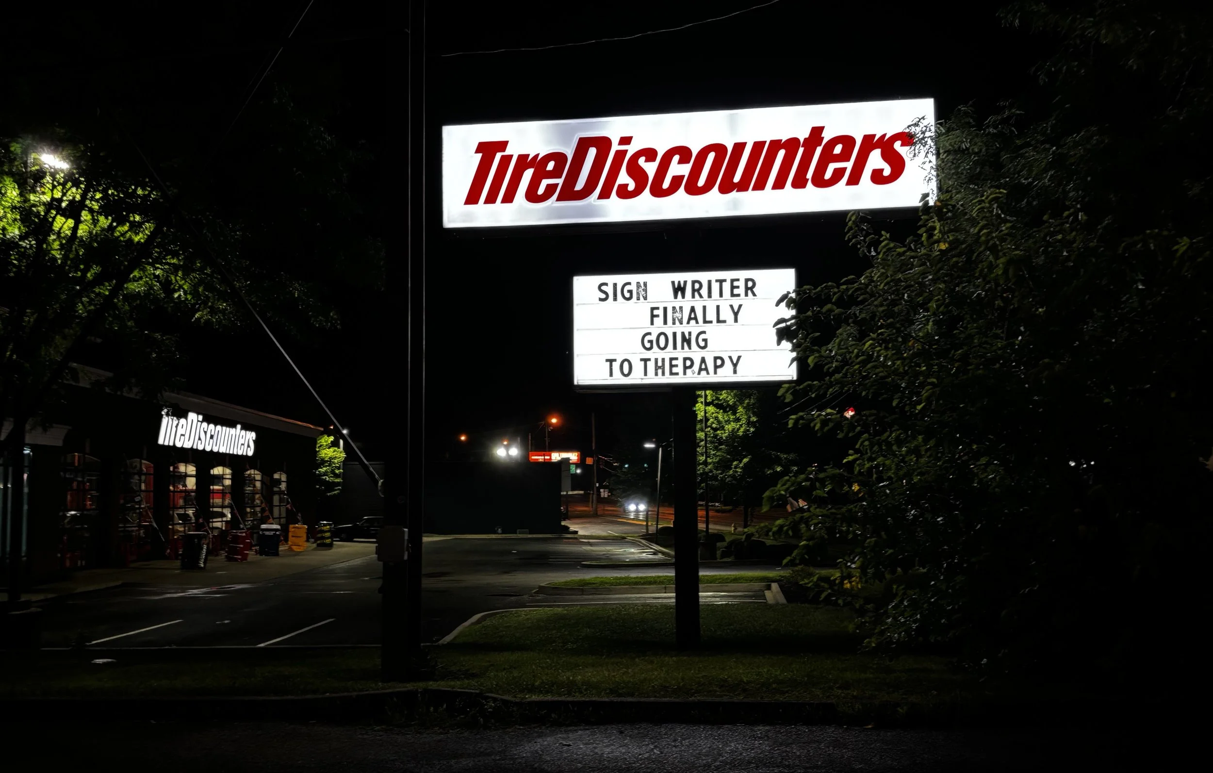 A brightly lit sign for Tire Discounters at night, with a humorous message on a smaller sign below that reads, 'SIGN WRITER FINALLY GOING TO THE PAPER.'