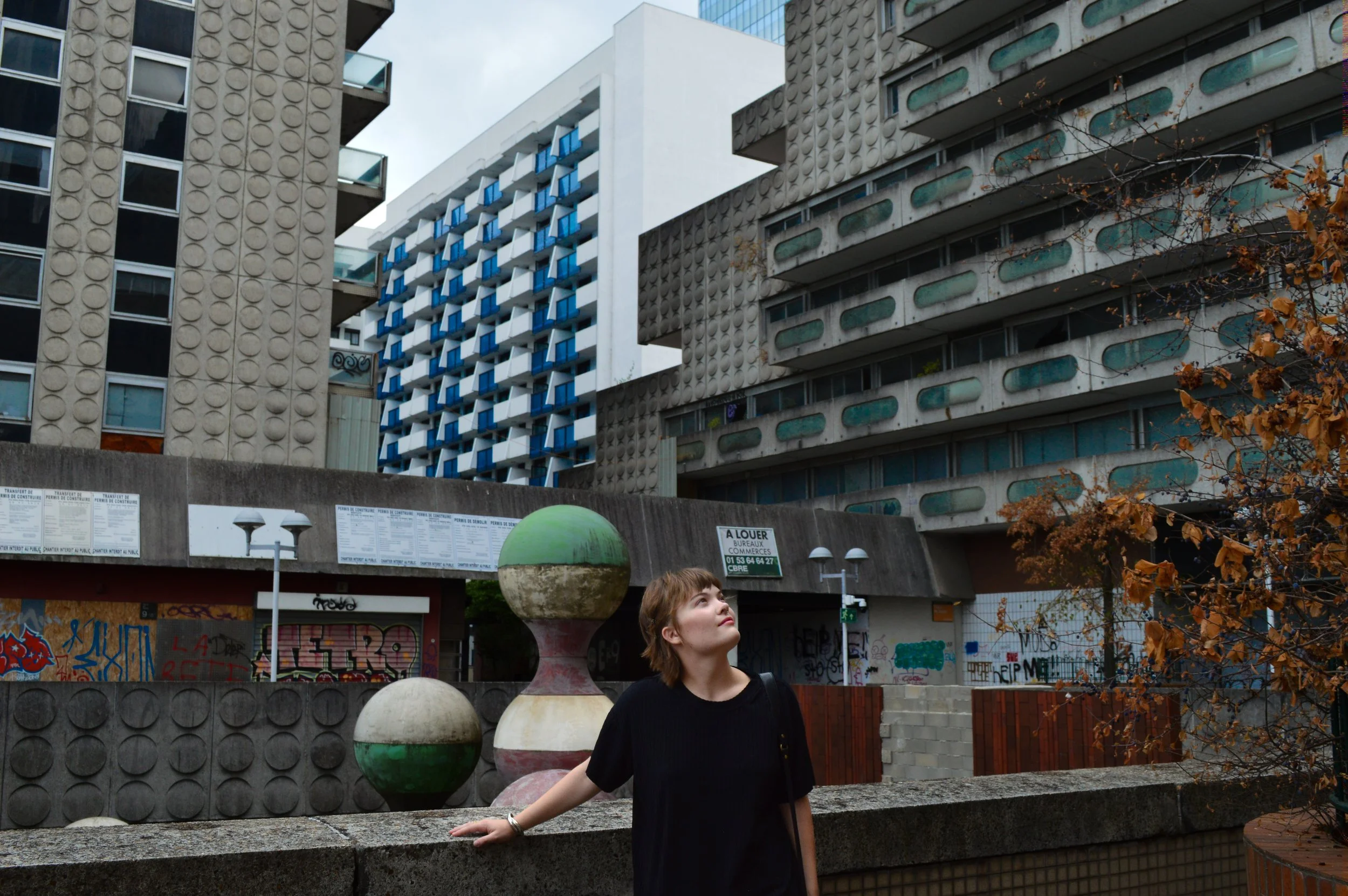A young woman with short brown hair wearing a black shirt standing outdoors in an urban area with modern buildings, graffiti-covered walls, and fallen autumn leaves. She is leaning against a stone ledge with her arm resting on it, looking upward. There are colorful decorative spheres on the ledge and graffiti on the walls behind her.