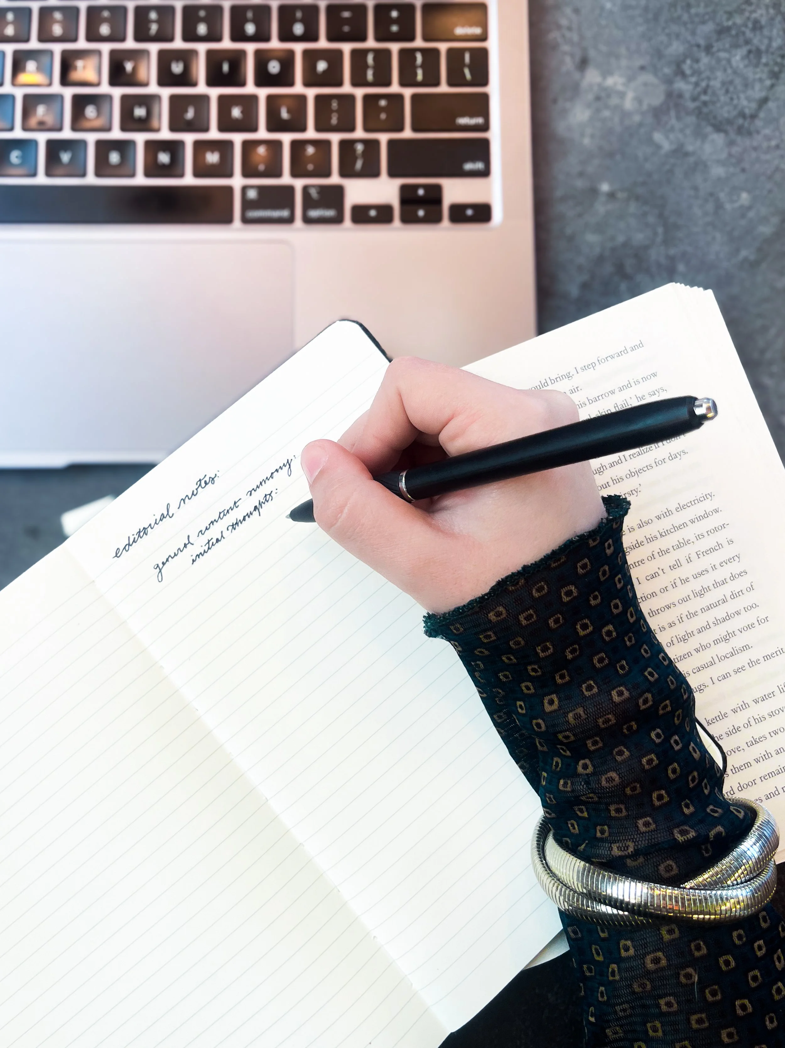 A person writing in a notebook with a black pen, wearing a dark patterned sleeve, near a silver laptop on a gray surface.
