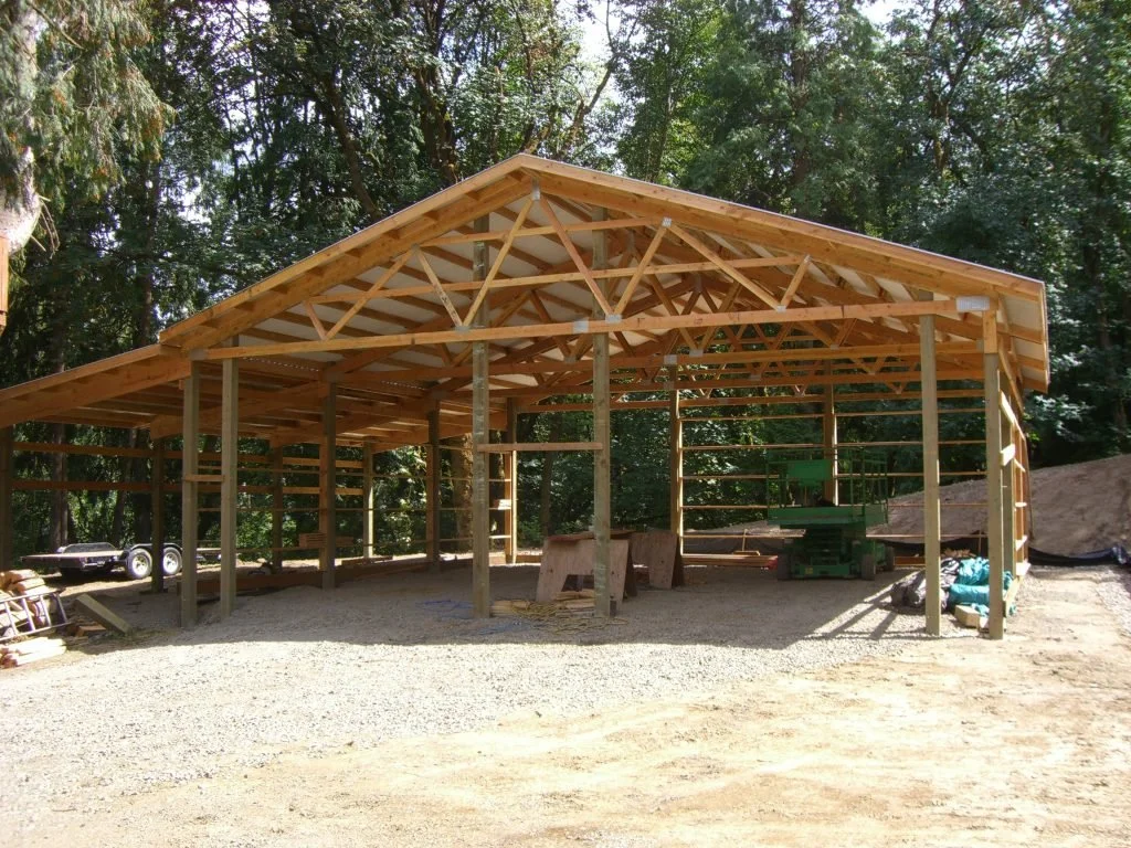 Wooden structure under construction with an open framework and a sloped roof, set outdoors surrounded by trees.