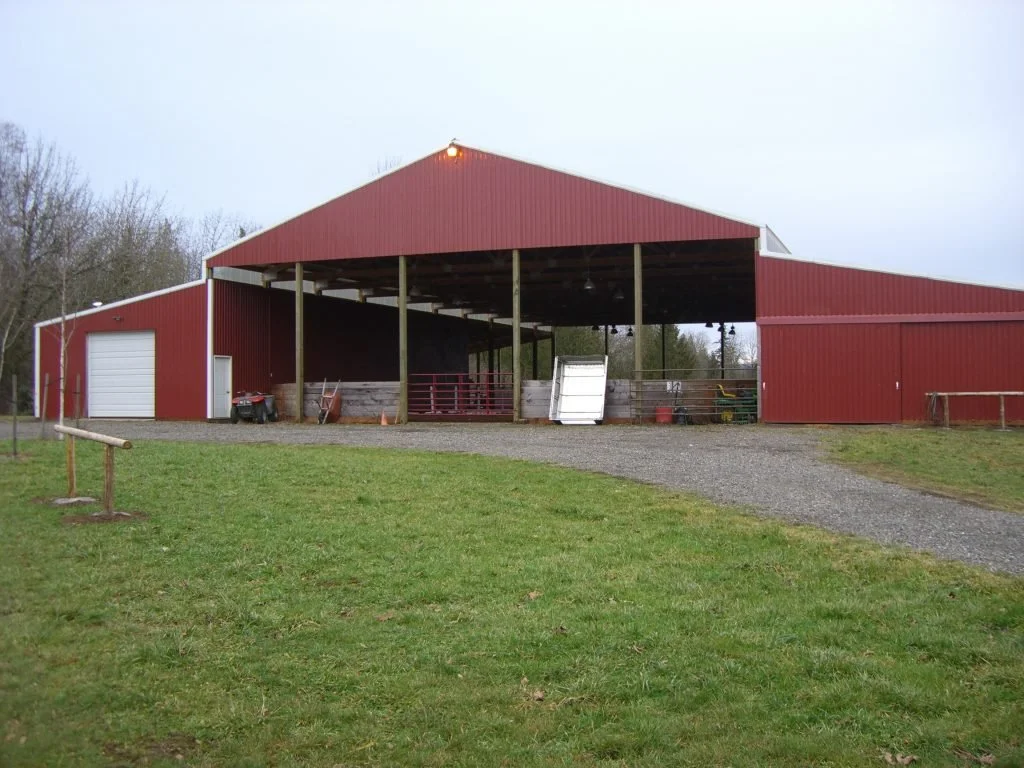 A red farm pole barn with open sections, a white garage door, and various farm equipment and supplies inside and outside.