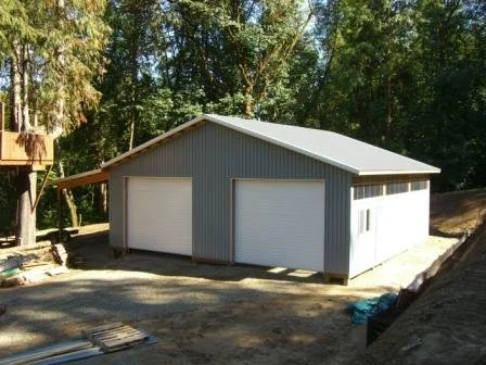 A gray metal garage with two roll-up doors on a concrete foundation, located in a wooded area.