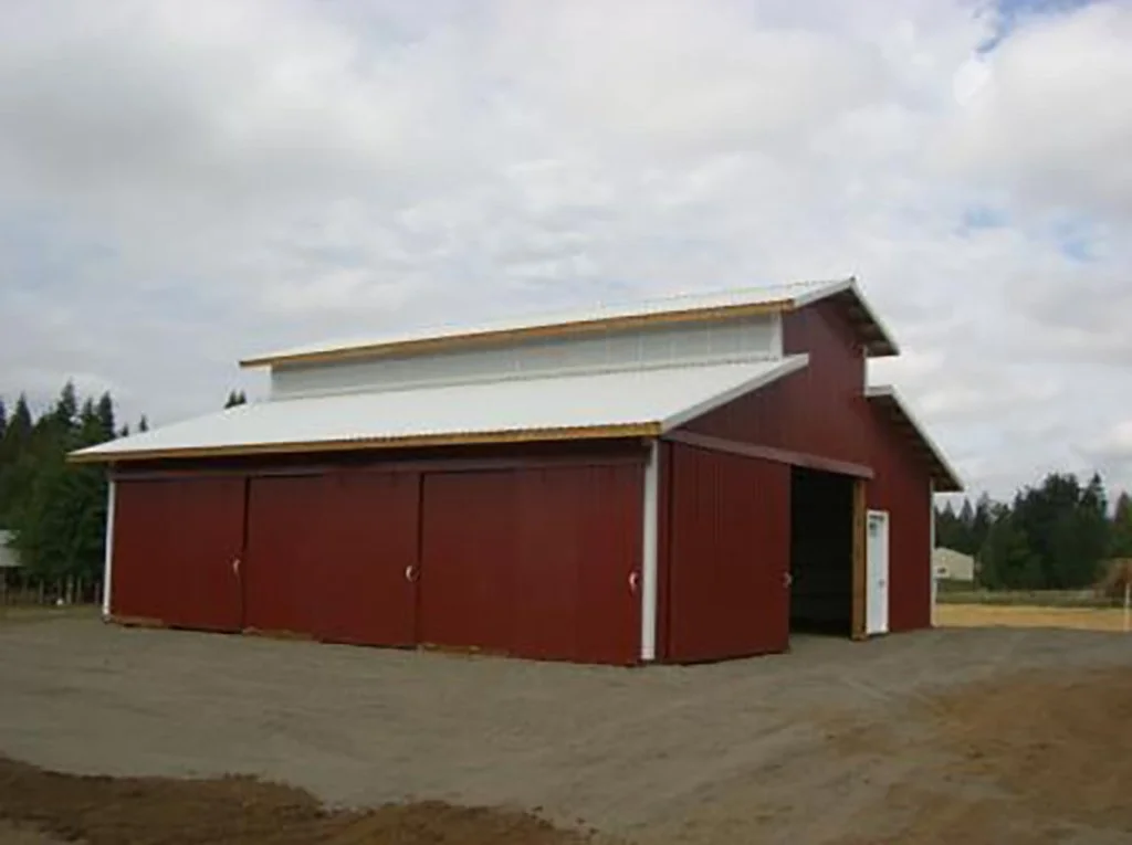 A red barn with a white roof on a gravel lot, surrounded by trees and cloudy sky.