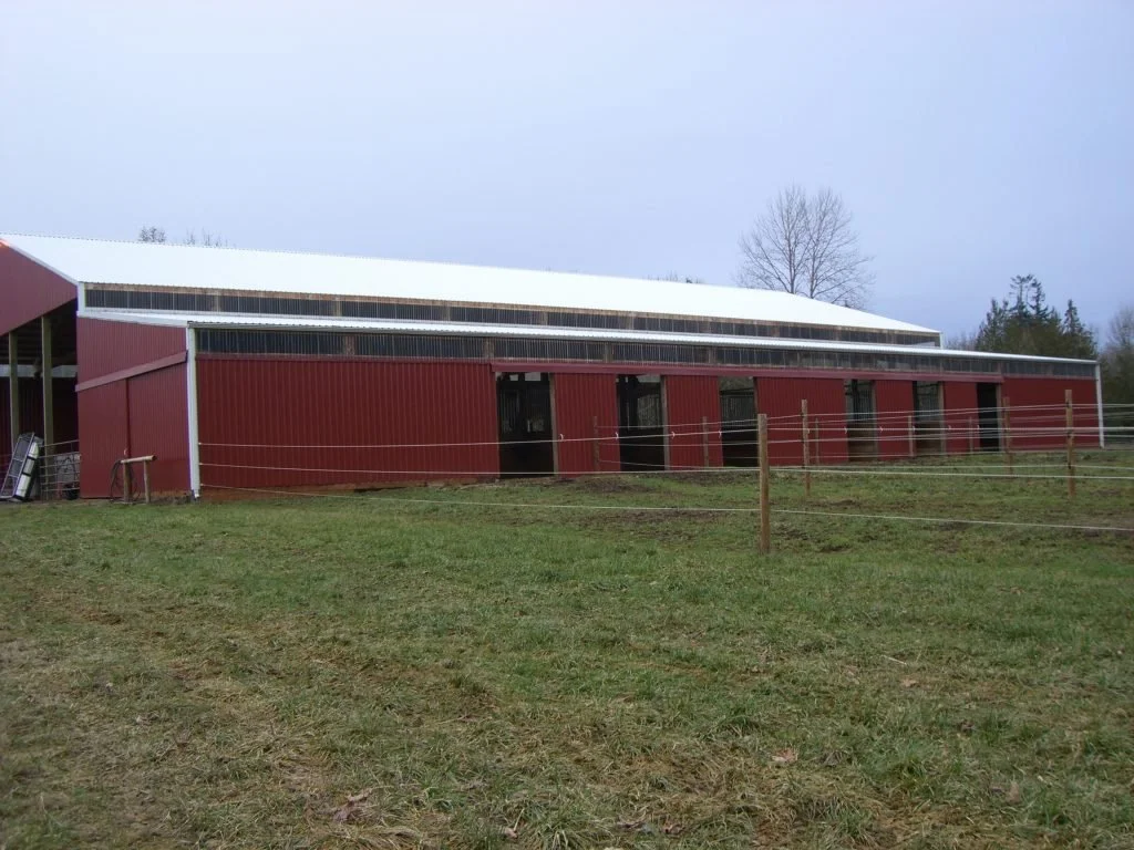 A large red barn with a white roof on a farm, enclosed by wooden fencing, with a grassy field in the foreground and trees in the background.