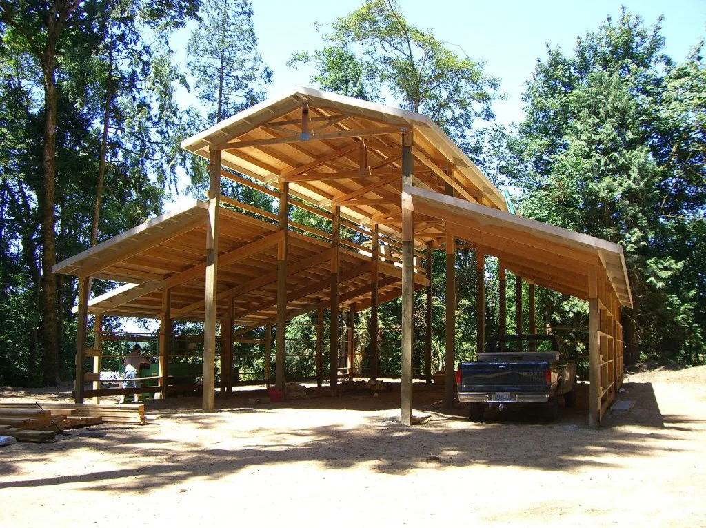 Wooden house under construction in a wooded area, with a truck parked in the garage, and workers on site.