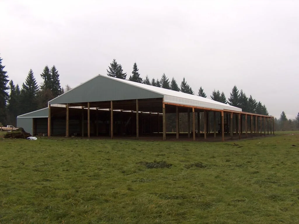 A large open-sided barn with a metal roof and wooden supports, situated on a grassy field with trees in the background.