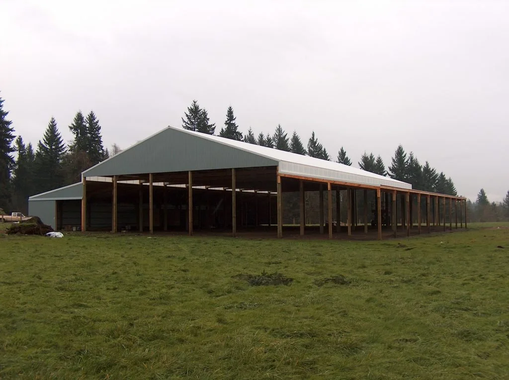 An unfinished large metal building with a gabled roof on a grassy field, surrounded by trees.