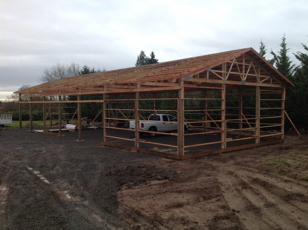 Wooden framework for a building under construction with a pickup truck inside. Dirt ground with tire tracks and darker soil in the foreground, trees in the background, overcast sky.