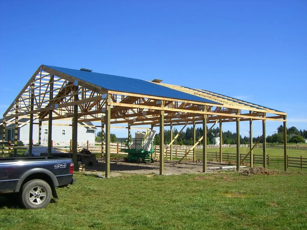 Wooden structure being built, possibly a garage or large shed, with a blue roof and construction equipment nearby, in a grassy area with a house and trees in the background.
