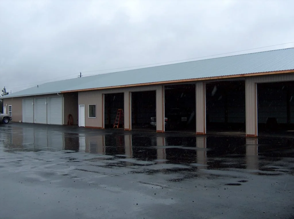 A large metal warehouse with open garage doors and a single closed door, rain-slicked asphalt in front, overcast sky, parked vehicles visible inside and outside.