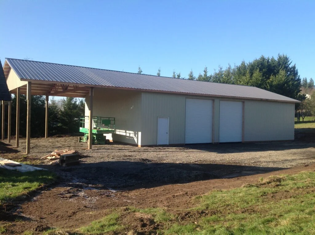 A large beige metal garage with two roll-up doors and one side door, situated on a mostly dirt lot with some grassy patches, under a clear blue sky.