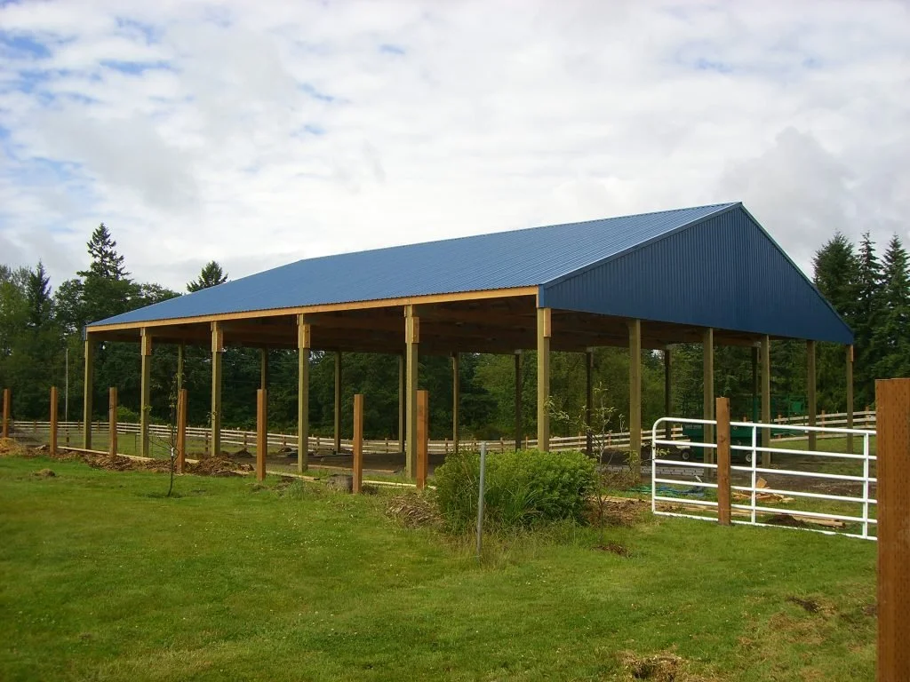 A partially built outdoor pavilion with a blue metal roof and wooden support beams, situated on a grassy area with trees in the background.