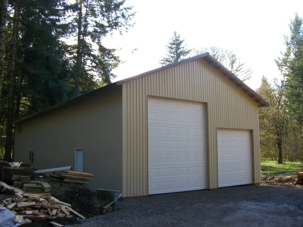 A metal building with two white garage doors and siding, situated in a wooded area with tall trees, under a partly cloudy sky.