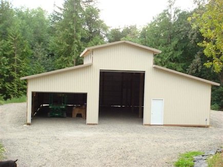 A large beige storage shed with a gable roof, open in the center, with two side sections, surrounded by trees and a gravel ground.