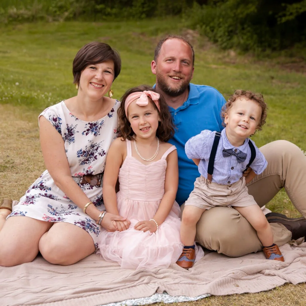 A family of four sitting on a blanket outdoors on a grassy field, smiling at the camera. The mother has short dark hair, is wearing a white floral dress. The father has a beard, is wearing a blue polo shirt and tan pants. One girl has long brown hair, a pink dress and a pink bow headband. The boy has curly hair, a light blue shirt with suspenders and a bow tie, and tan shorts.