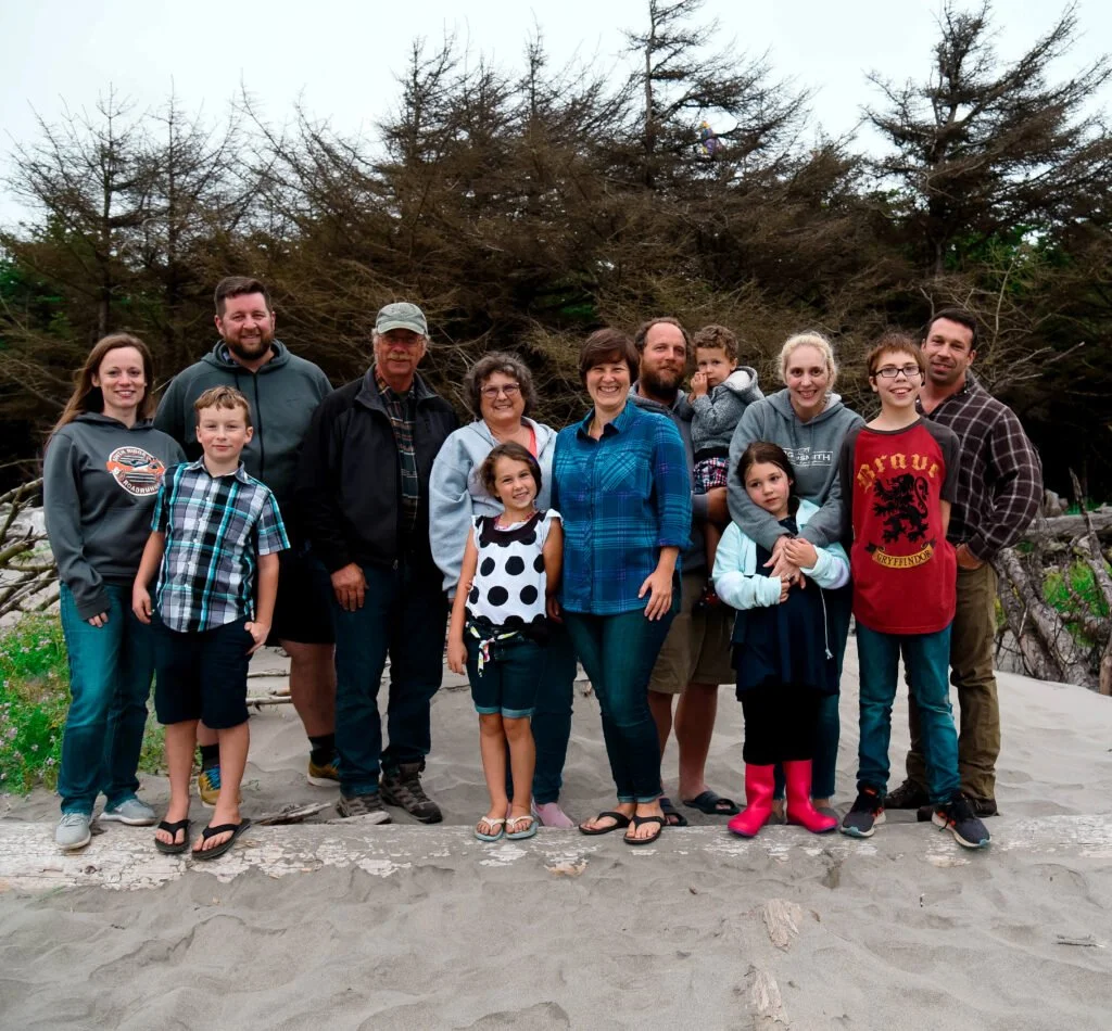 A group of twelve people, consisting of children and adults, standing on a sandy beach with driftwood and large evergreen trees in the background.