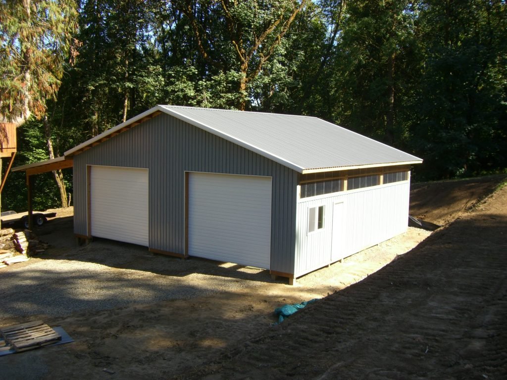 A newly built metal garage with two roll-up doors, situated on a dirt lot surrounded by trees.